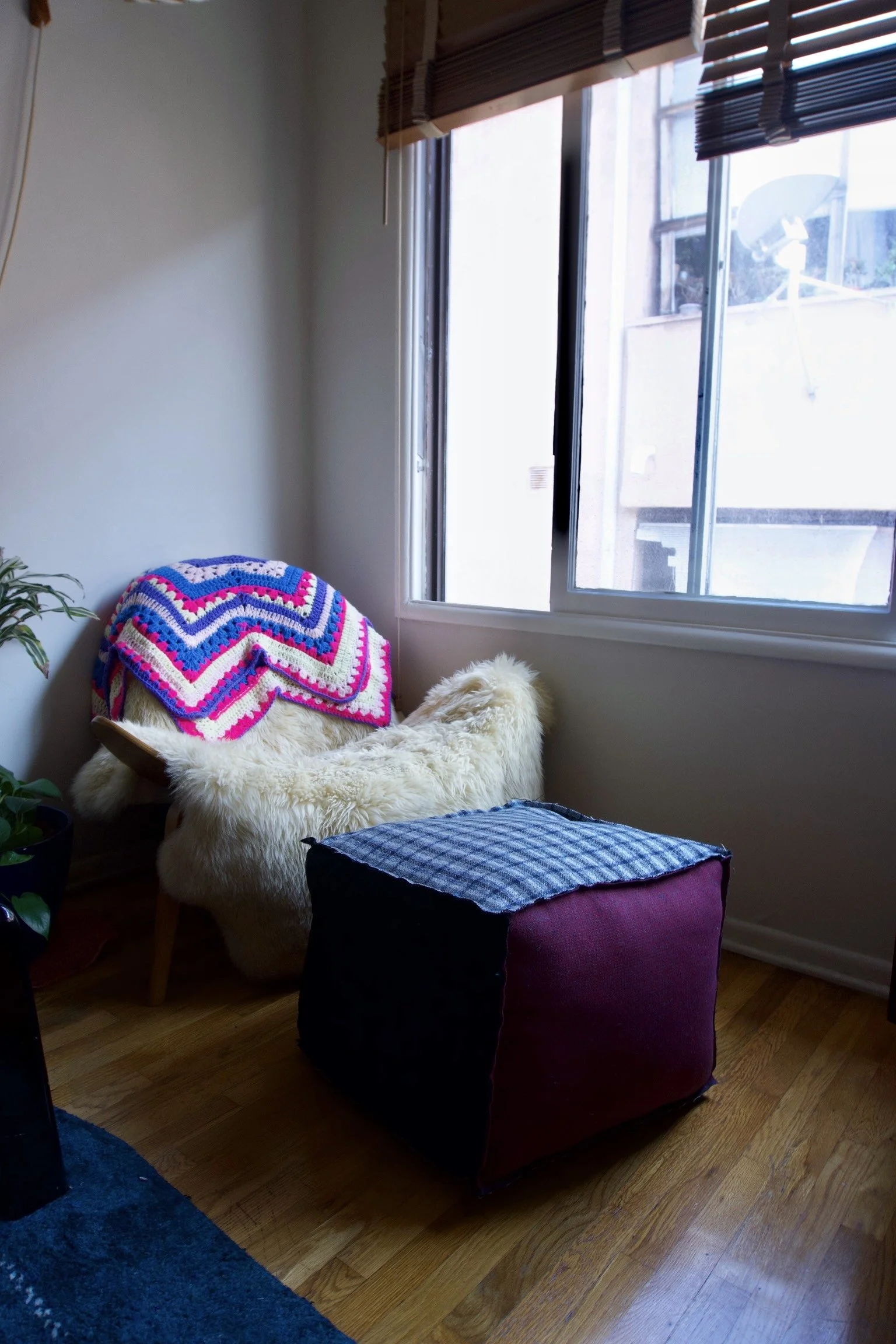 A cozy corner with a plush armchair covered with a cream-colored furry throw, accented by a colorful crochet blanket, beside a window with wooden blinds. A fabric-covered ottoman in front of the chair and a green potted plant nearby suggest a comfort