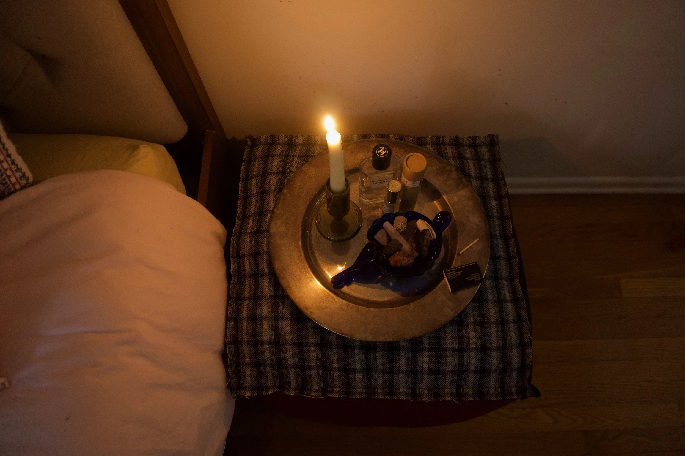 A bedside table with a lit candle, various bottles, and a bowl of crystals or stones, next to a bed with a beige pillow and a patterned pillow in a dimly lit room.