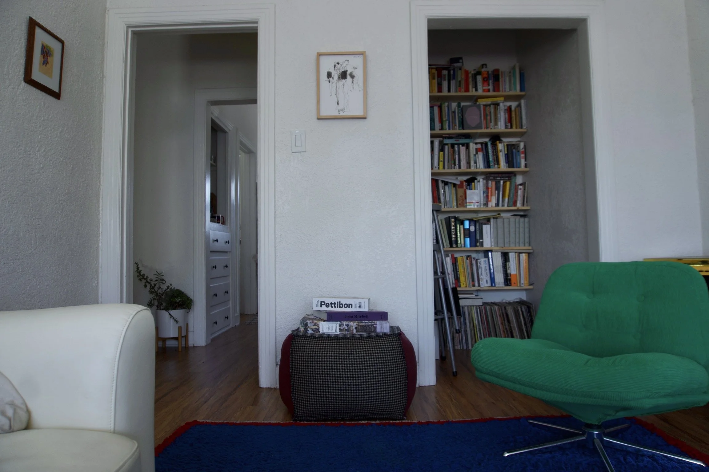 Living room corner with a white armchair partially visible on the left, a green lounge chair on the right, a small table with books and magazines in the center, a blue rug on the floor, a bookshelf in an alcove to the right, and framed artwork on the