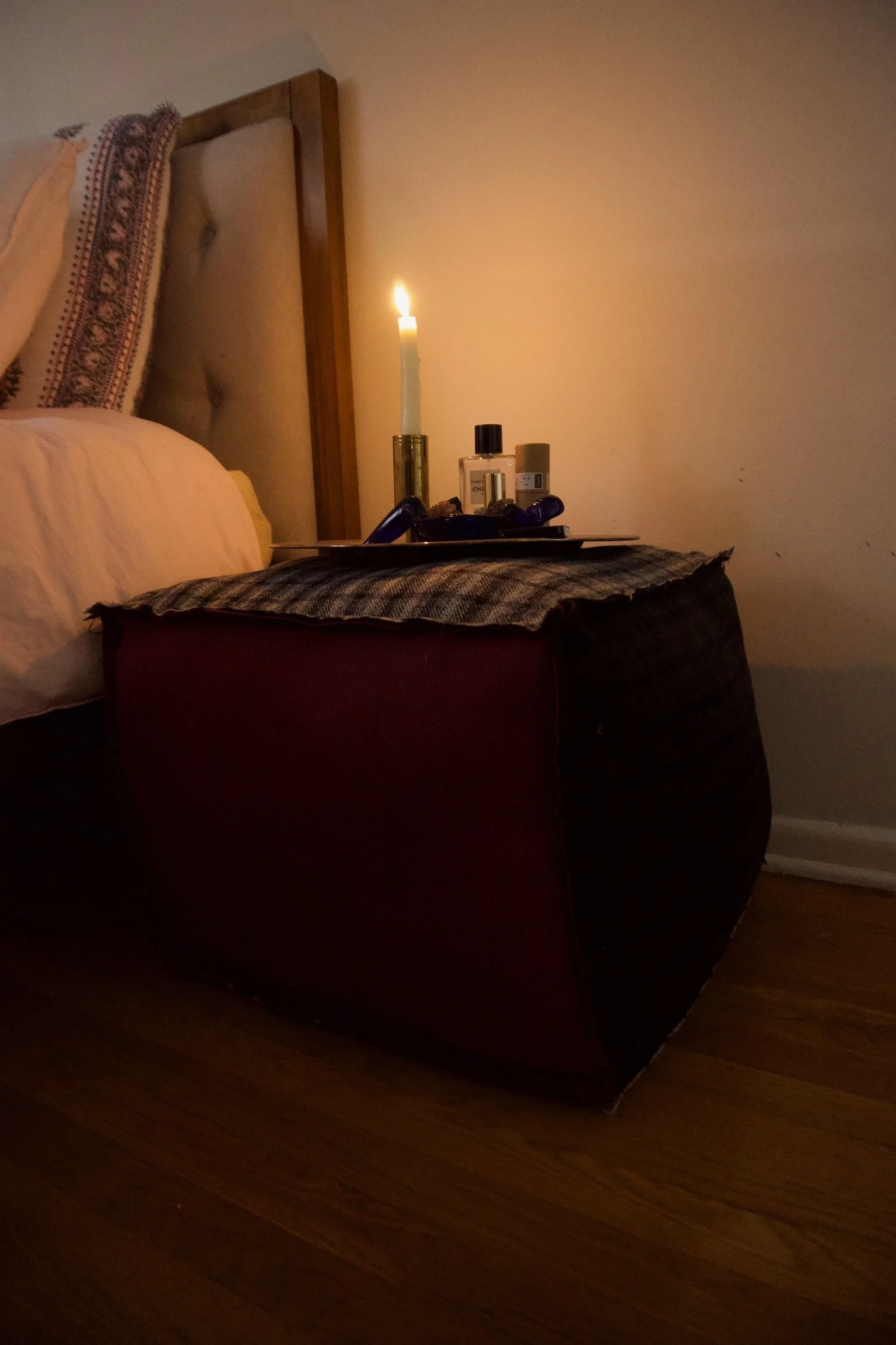 A bedroom nightstand holding a tray with perfume bottles, a candle in a holder, and other small items, with a bed and pillows in the background. The candle is lit, casting a warm glow.