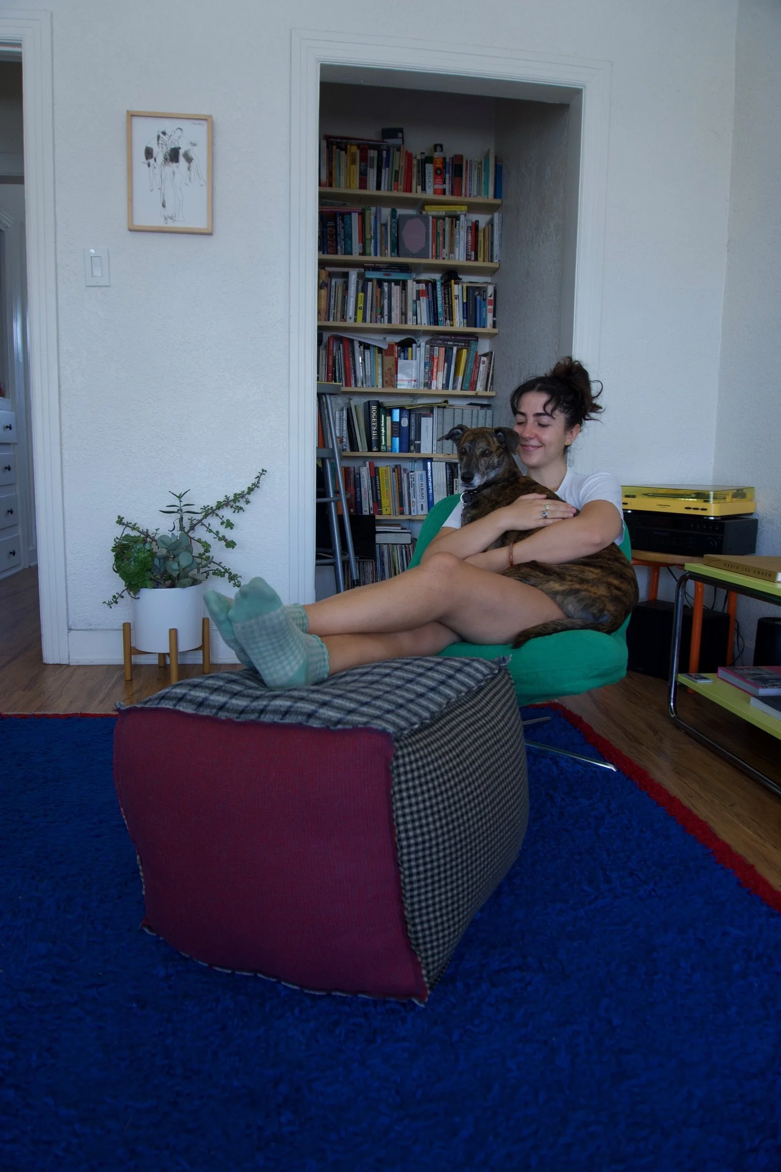 A woman sitting in a green chair with her dog on her lap in a cozy living room with a blue rug, a black and red ottoman, a potted plant, a bookshelf, and a yellow record player.