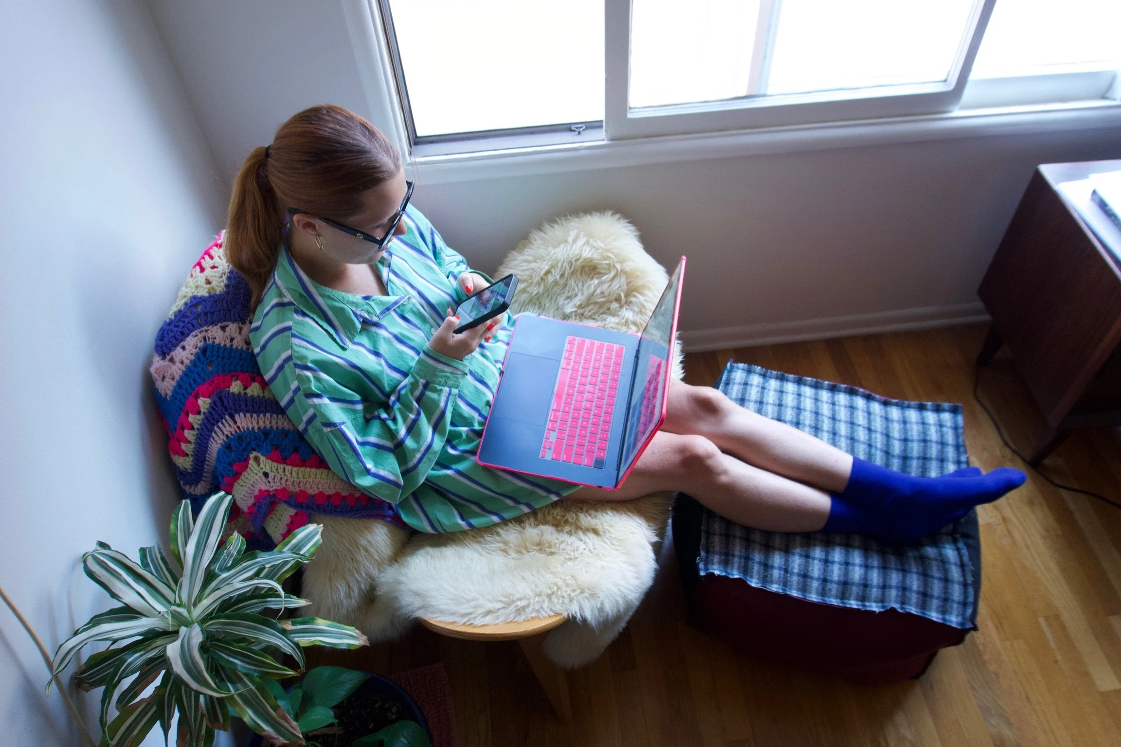 Young woman with glasses, in striped pajamas, sitting on a cozy chair with a pink laptop and smartphone, with her feet on an ottoman covered with a checked cloth, near a window with natural light, a plant nearby, and a colorful crocheted blanket behi