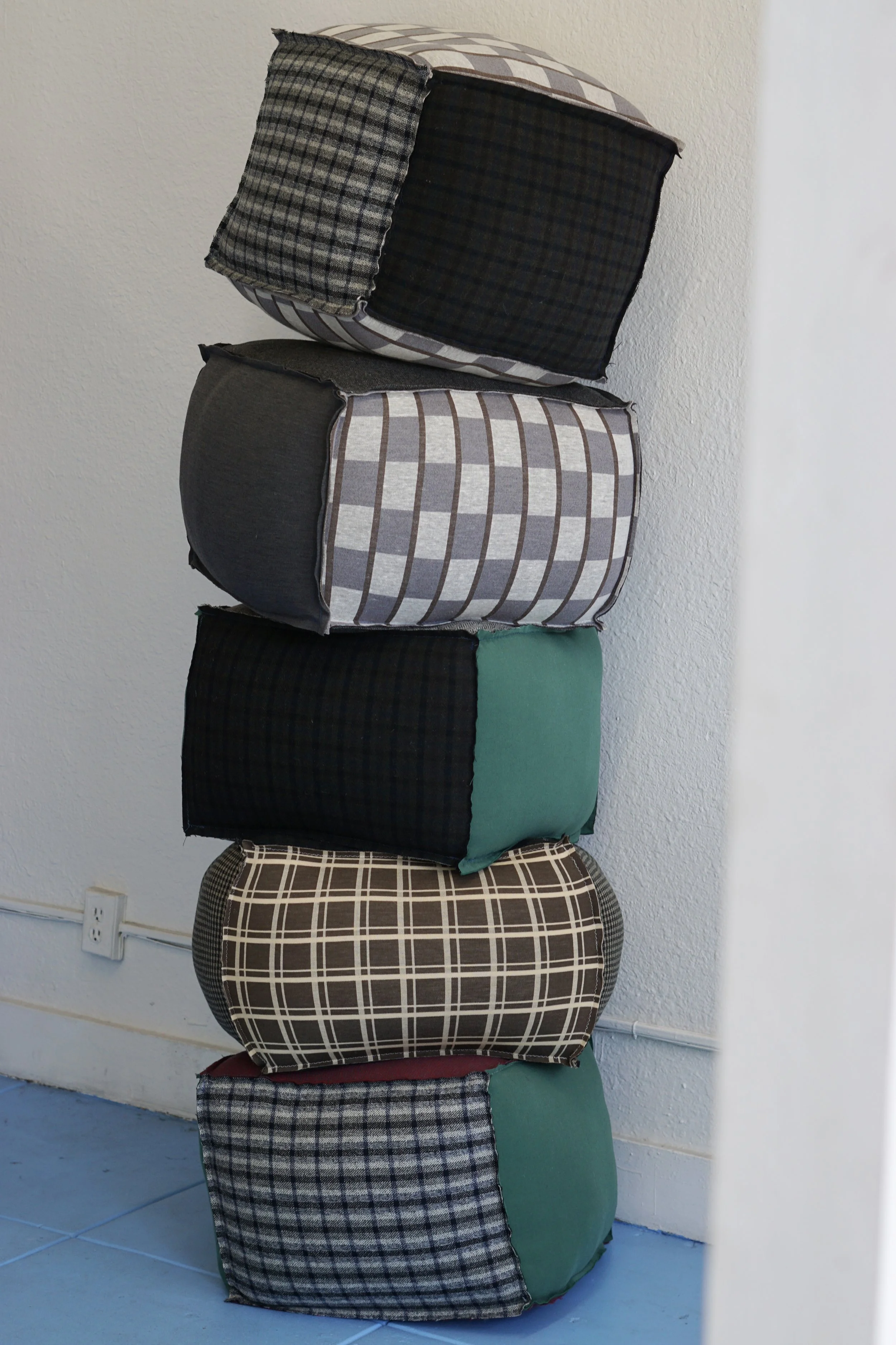 A stack of five cube-shaped cushions with various plaid and checkered fabric patterns, stacked vertically against a white wall next to a power outlet.
