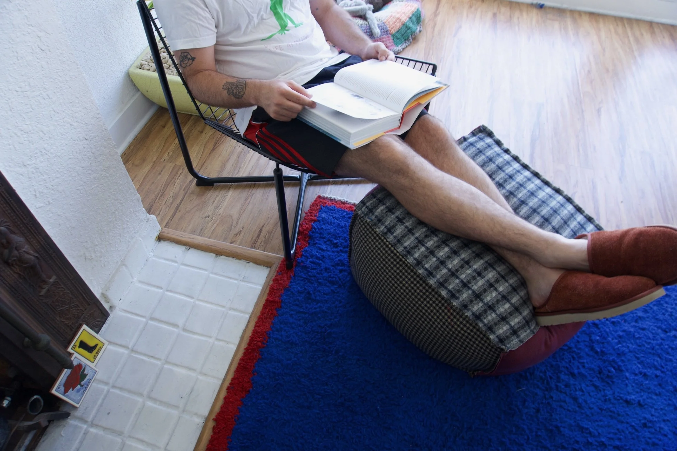 Person sitting on a black wire chair reading a large book, with feet resting on an ottoman, wearing slippers in a room with hardwood floor and a colorful rug.