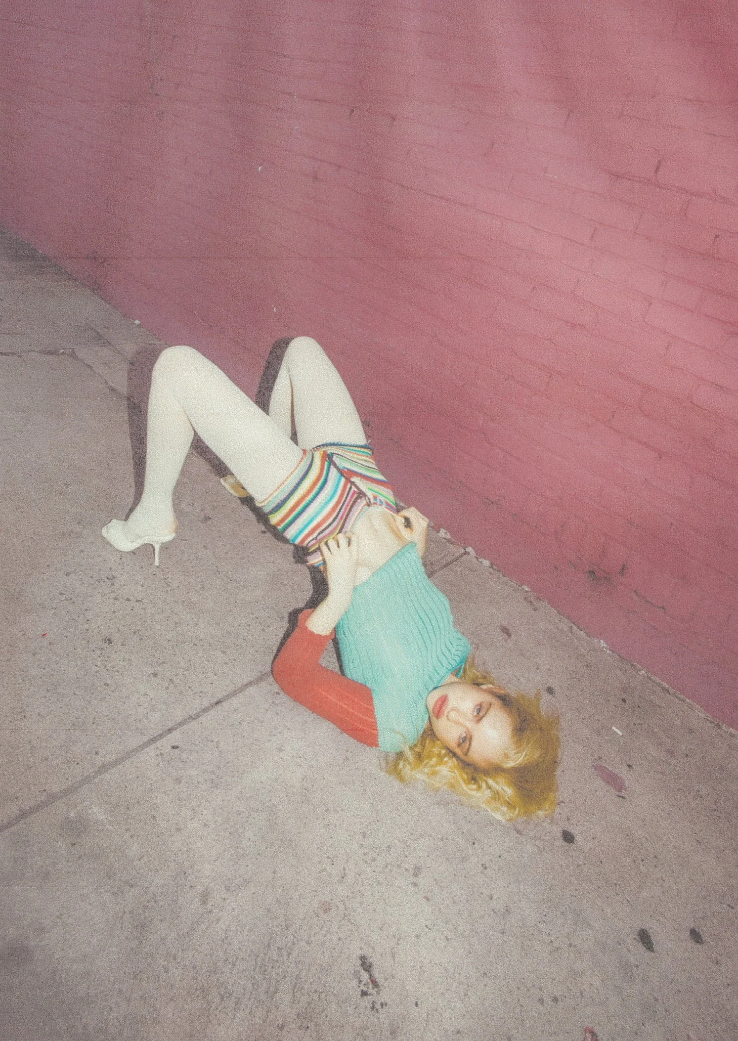 A woman with curly blonde hair, wearing a teal and orange long-sleeve shirt, a multicolored striped skirt, white tights, and white heeled shoes, lying on her back on a concrete sidewalk next to a pink brick wall, looking at the camera.