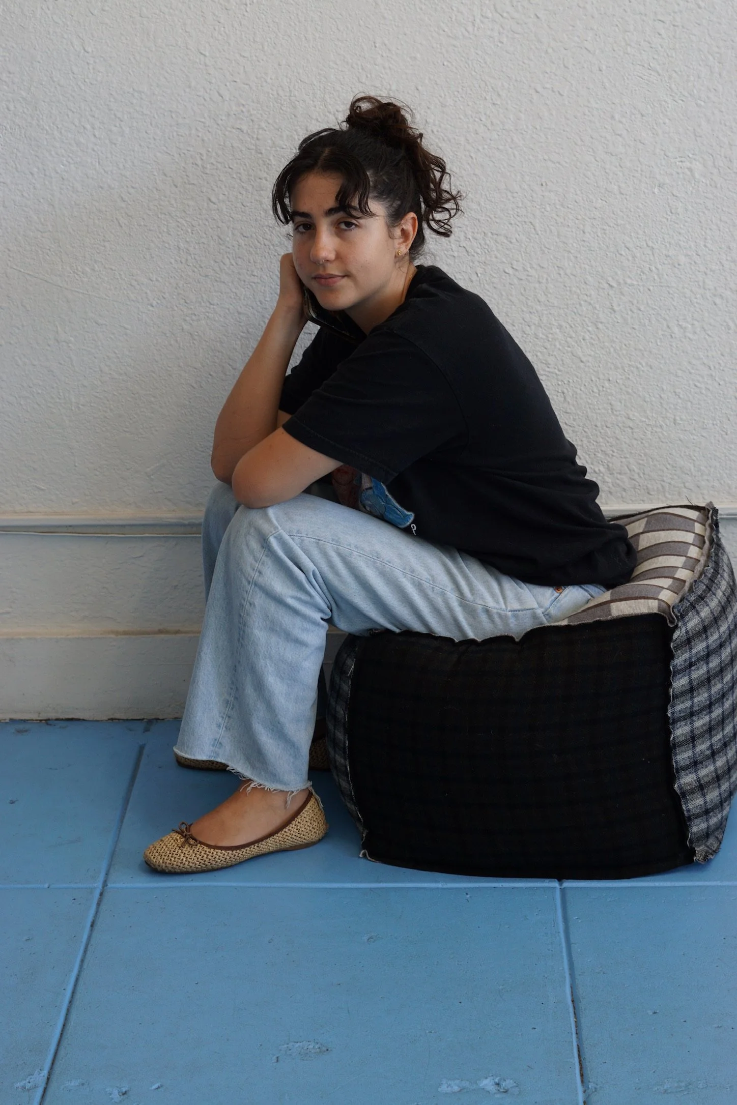 Young woman with dark hair in a bun, sitting on a checked pillow on blue tile floor, resting her head on her hand, wearing a black t-shirt, light blue jeans, and beige flats, against a white textured wall.