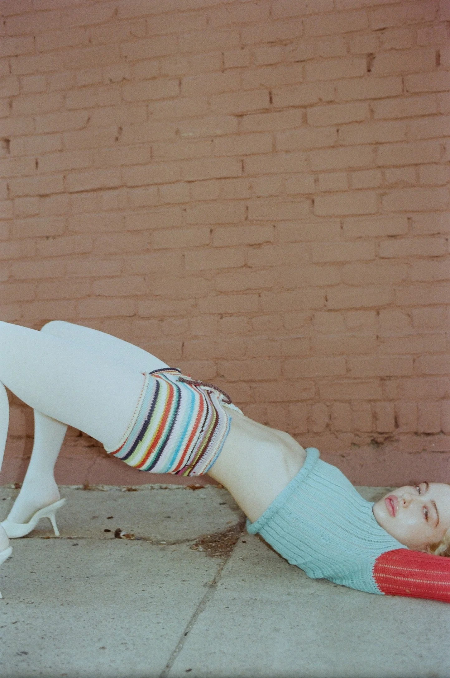 A woman lying on the ground with her torso arched back, wearing a colorful sweater, white tights, a striped sock, and high heels, against a brick wall.