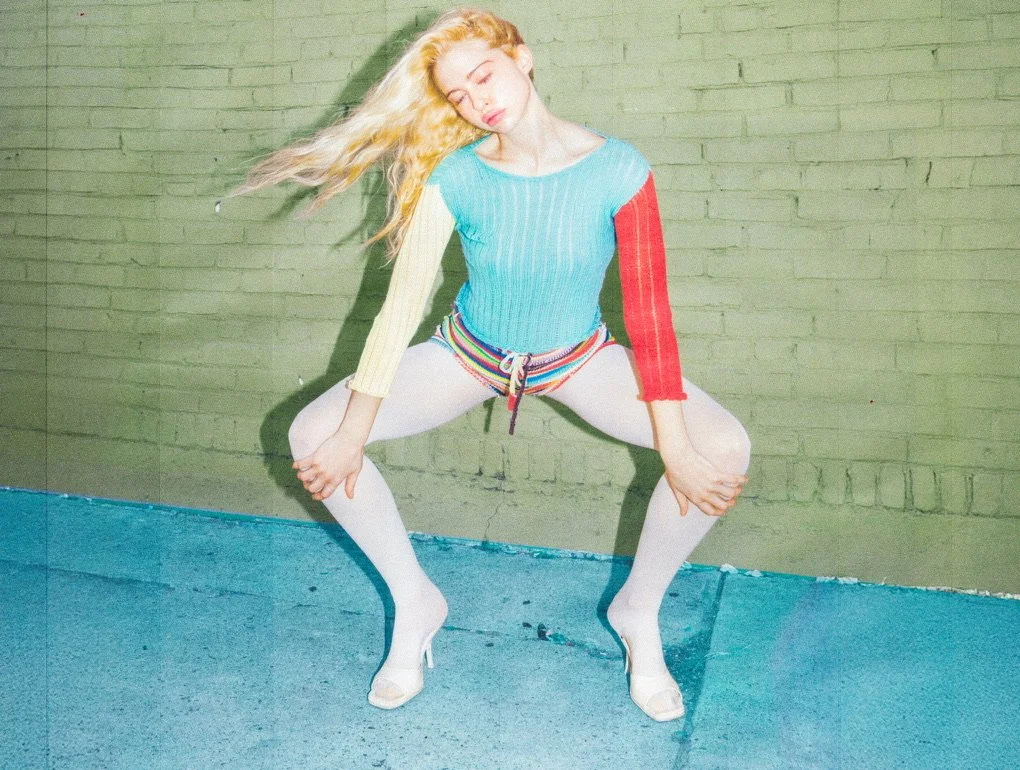 A young woman with long blonde hair posing against a green brick wall, wearing a colorful sweater, white leggings, and high heels, with her hands on her knees and eyes closed.