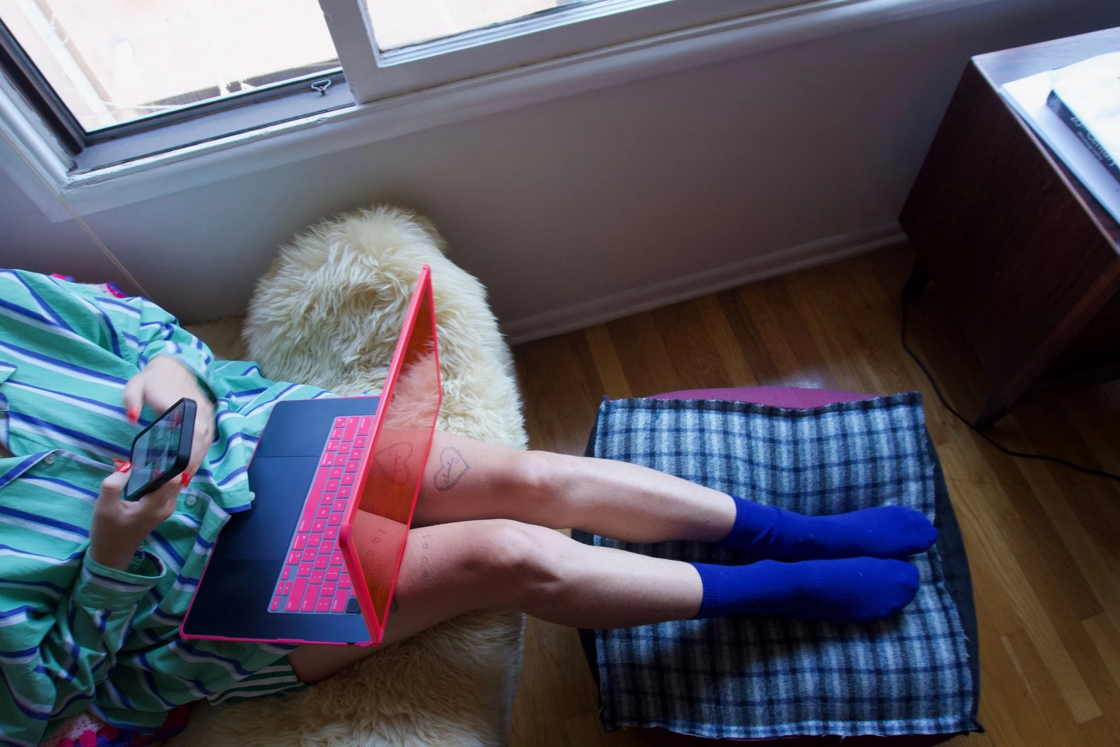Person lying on a bed with a pink laptop on their lap, holding a phone in hand, wearing blue socks and a striped shirt, next to a fluffy cream-colored dog.