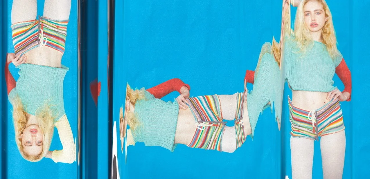 A woman with blonde hair standing in front of a mirror, wearing a rainbow striped crop top and matching shorts, with her reflection visible. The image is distorted by the mirror, creating a wavy effect.