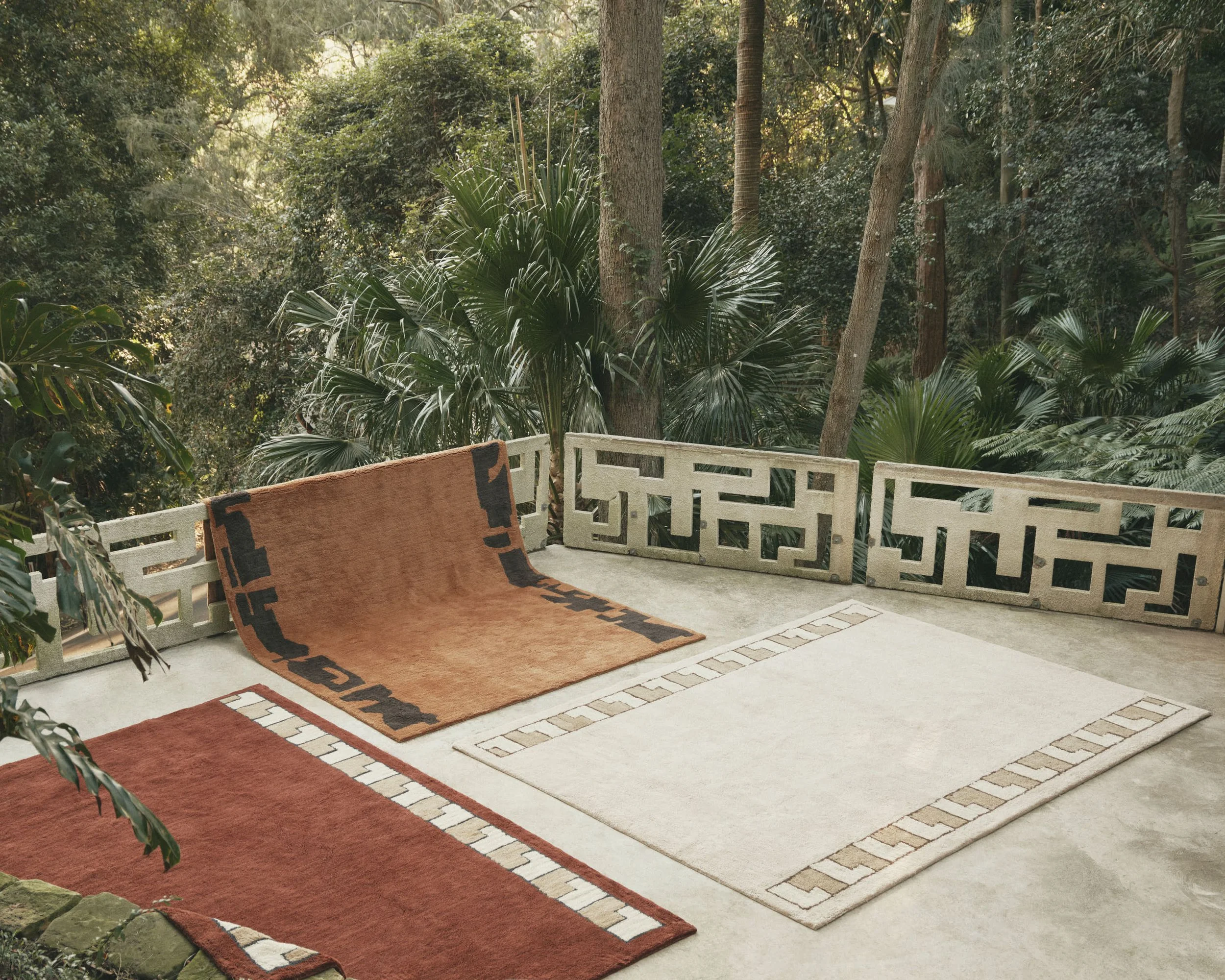 A terrace with two carpets laid out on a concrete floor, surrounded by lush green trees and plants, with part of a bench visible in the foreground.