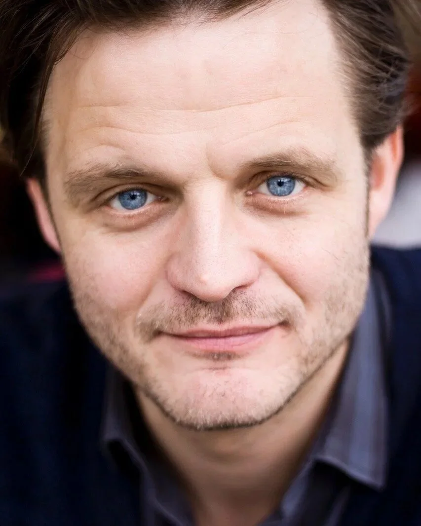 Nahaufnahme eines Mannes mit blauen Augen und braunem Haar, lächelnd, im Freien.