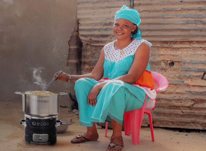 A woman sitting on a pink plastic chair, cooking on a portable stove outdoors, smiling at the camera, with a corrugated metal wall in the background.