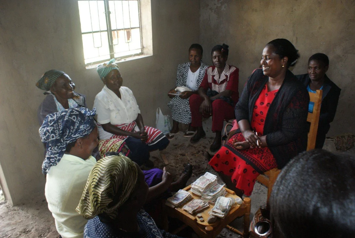A group of women sitting in a room, with one woman in a red dress and black cardigan smiling, surrounded by other women, some holding notebooks and money on a small wooden table.