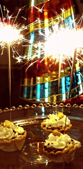 Mini dessert tarts topped with whipped cream and sparklers, displayed at a luxury celebration with warm ambient lighting in the background.
