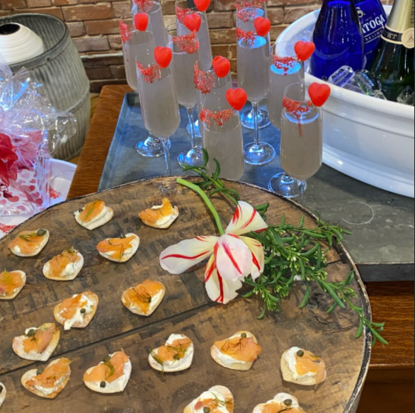Valentines Day party spread with heart-shaped salmon appetizers on wooden board and champagne cocktails garnished with strawberry hearts and pink sugar rim