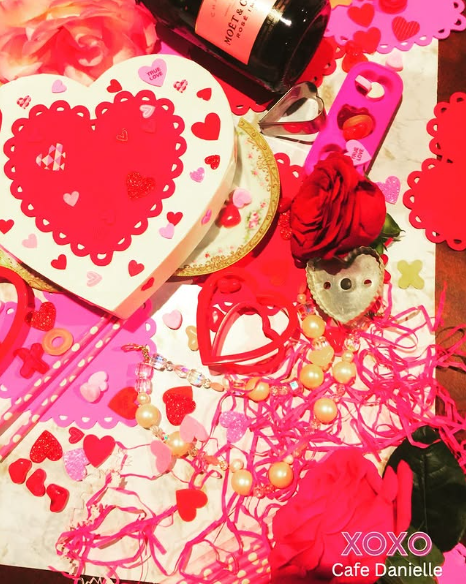 Valentines Day gift arrangement with heart-shaped boxes, red roses, champagne bottle, pearl jewelry, and romantic decorations on pink table at Cafe Danielle