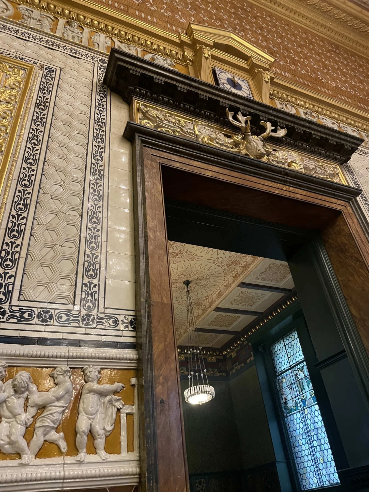  Decorative doorway framed by patterned tilework and gold detailing inside the Victoria and Albert Museum, with a stained-glass window and chandelier visible beyond. 