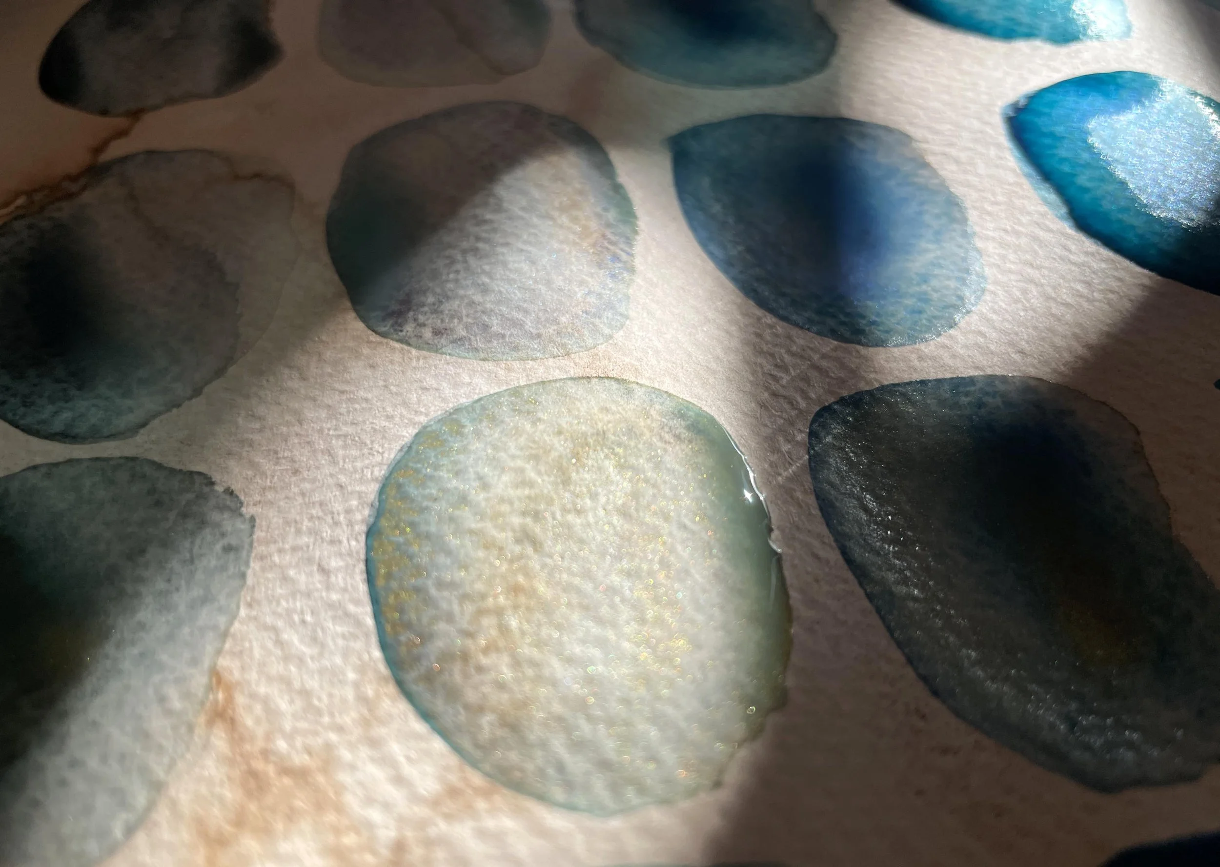 Watercolor color studies in blue and teal tones on textured paper, photographed in slanted sunlight with soft shadow.