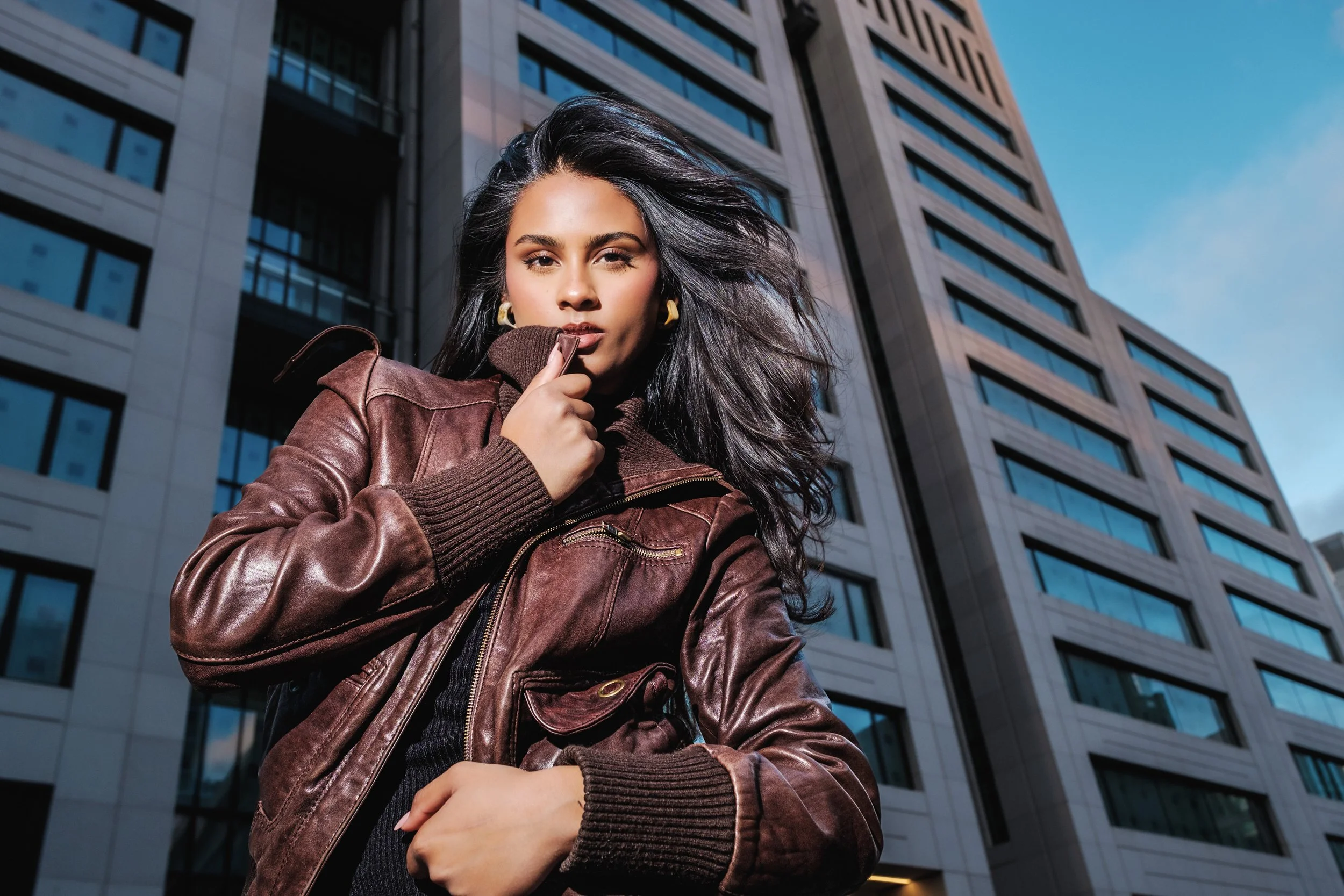 A woman with flowing dark hair wearing a brown leather bomber jacket poses in front of a modern high-rise building.