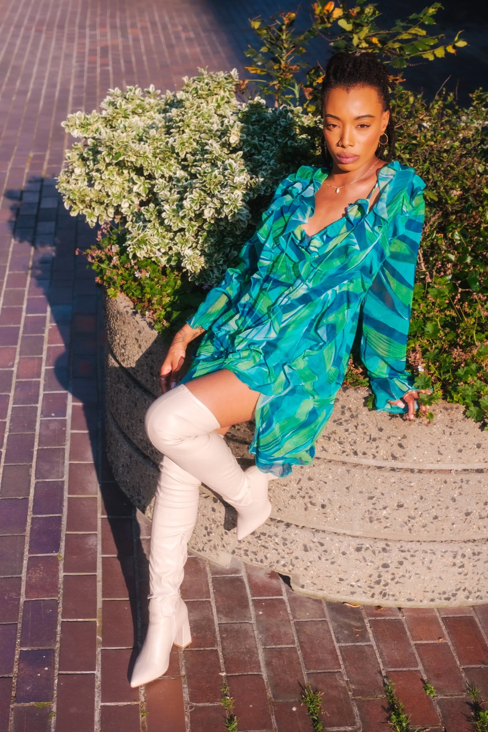 A woman with dark curly hair styled in a high ponytail wearing a vibrant blue and green patterned dress with ruffles, over-the-knee white boots, sitting against a circular concrete planter with lush green and white variegated plants, on a brick sidewalk during daytime.
