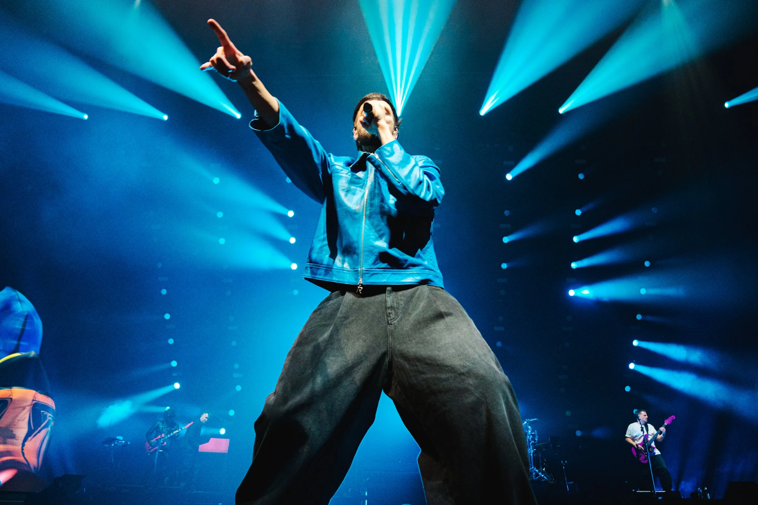 Kasabian lead singer Serge Pizzorno performs on stage in a blue leather jacket under bright teal spotlights at the O2 Arena