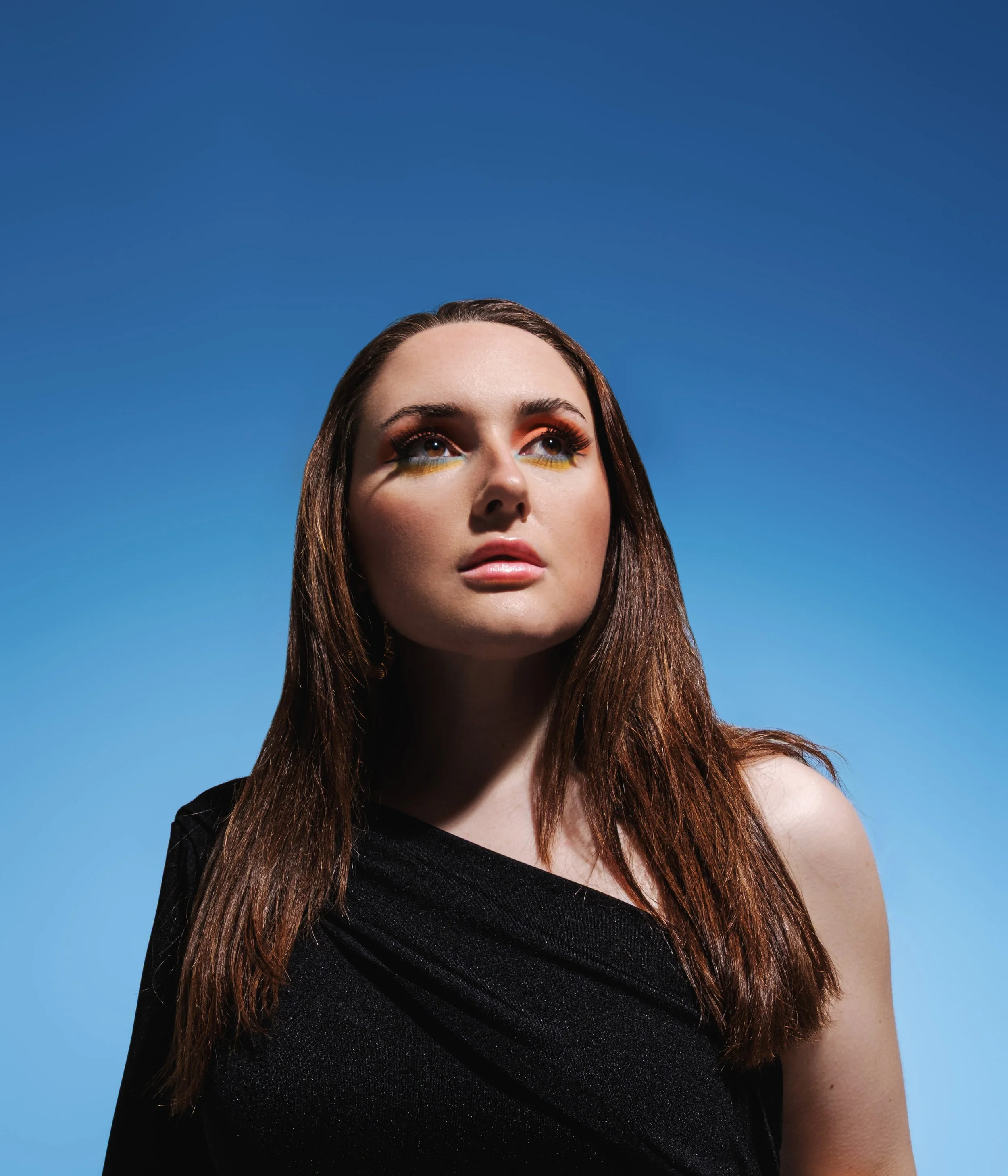 Editorial beauty portrait by Metty Unuabona featuring a woman with vivid orange and yellow eye makeup against a bold blue background.