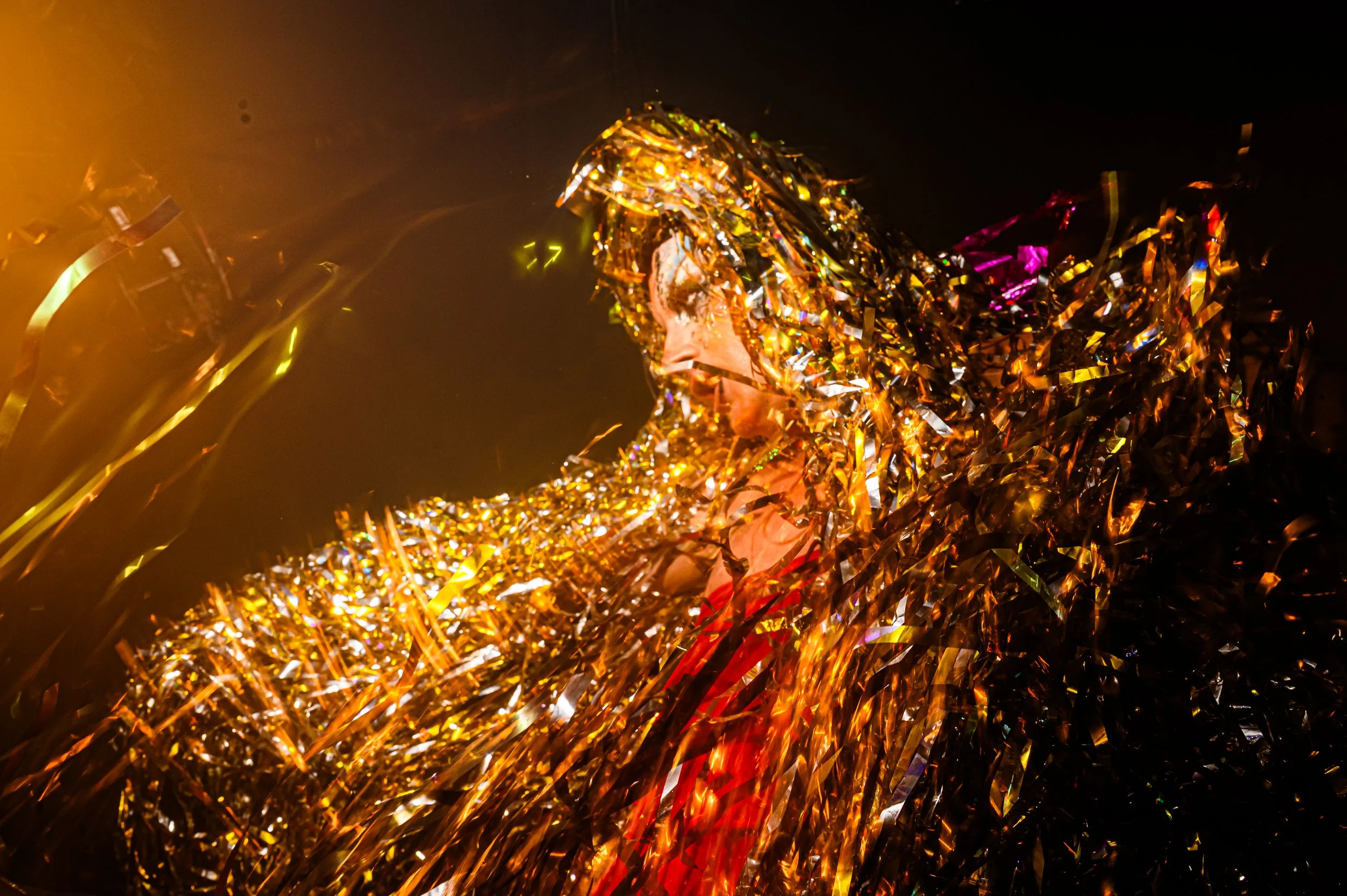 A performer at NYC Downlow Glastonbury covered in shimmering gold tinsel under dramatic stage lighting.