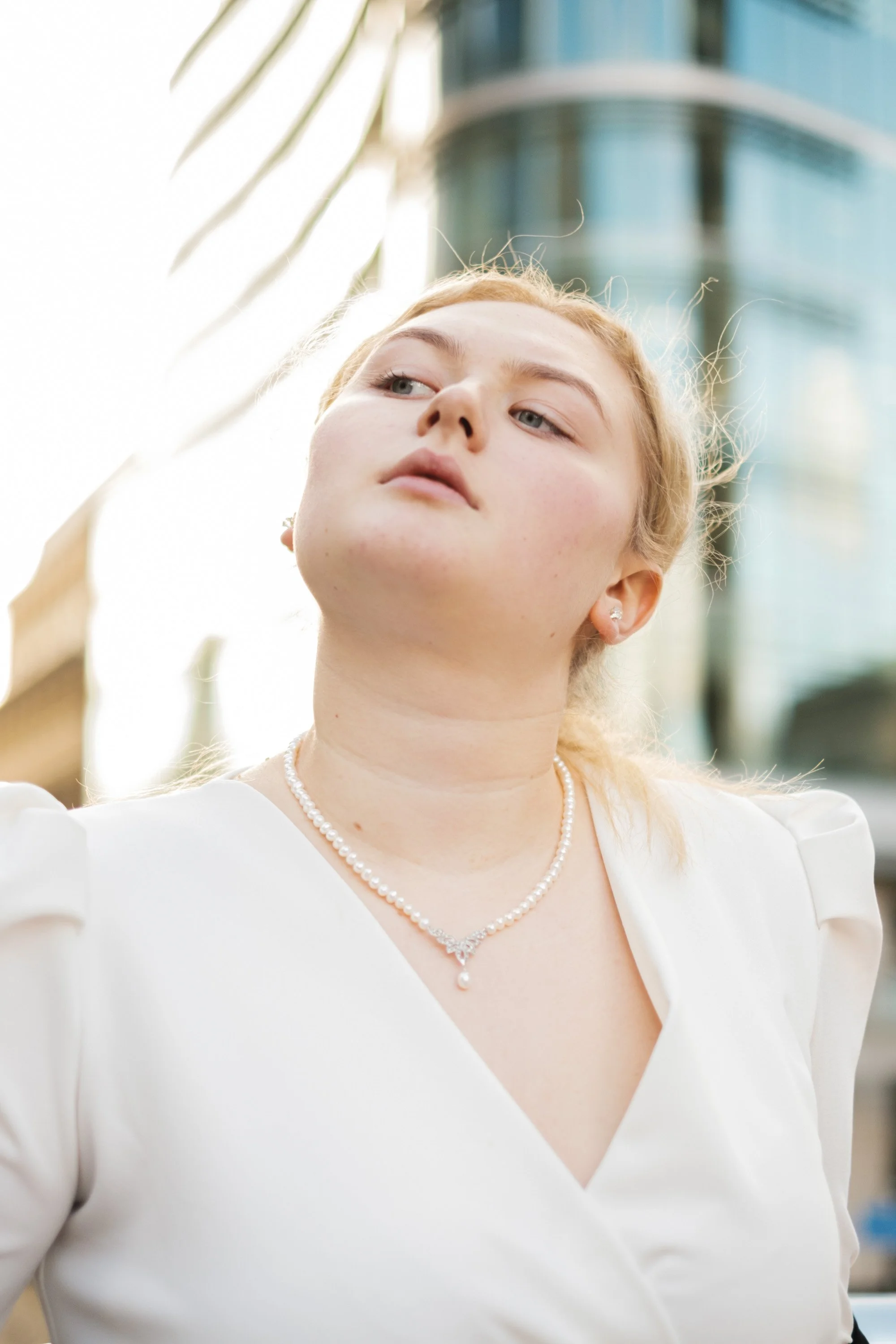 A young woman with fair skin and red hair wearing a white dress and pearl necklace, looking upward with a modern building in the background.