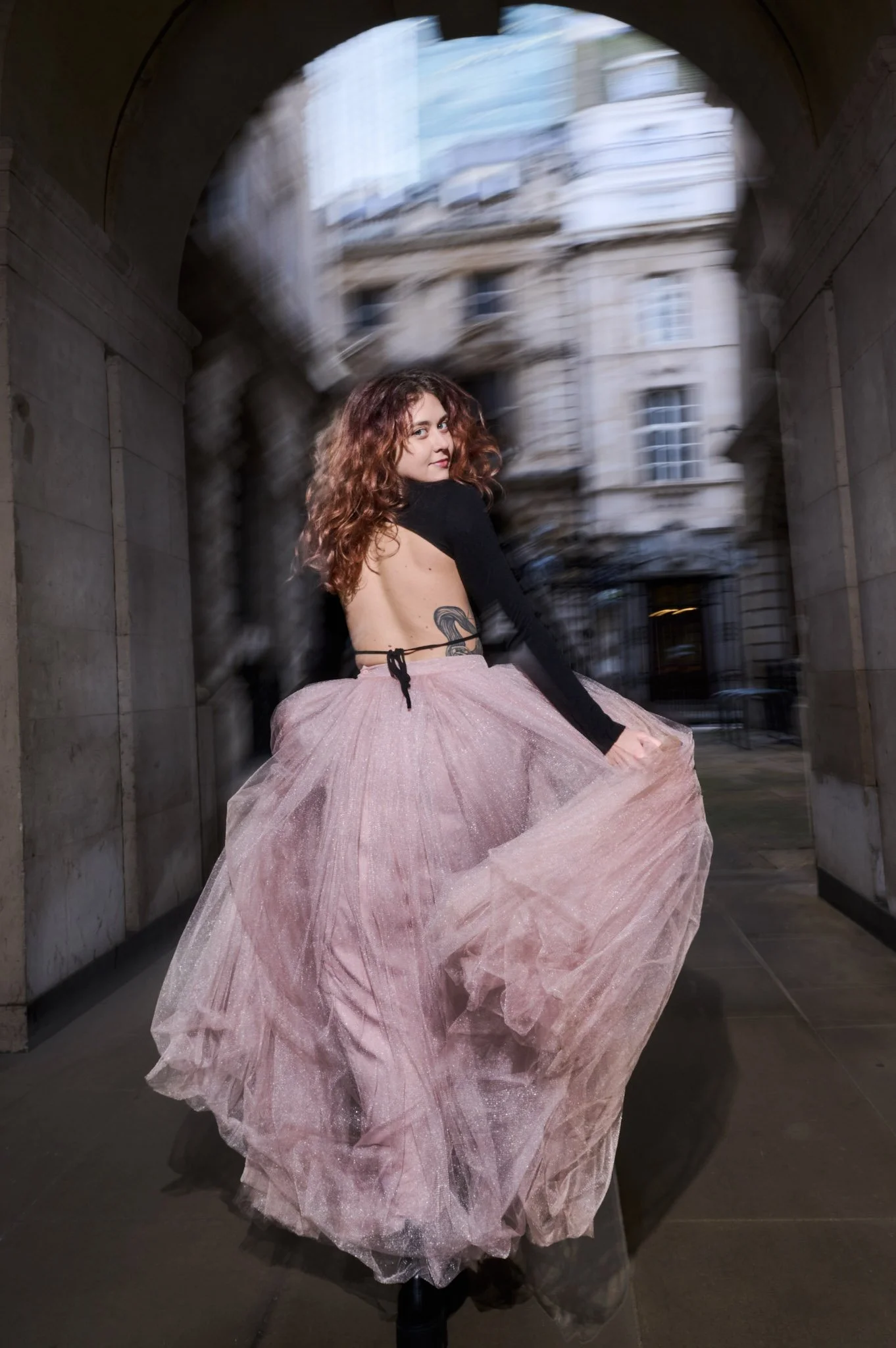 Young woman with curly hair wearing a pink tulle skirt and a black top with open back, standing under an archway in an urban setting.