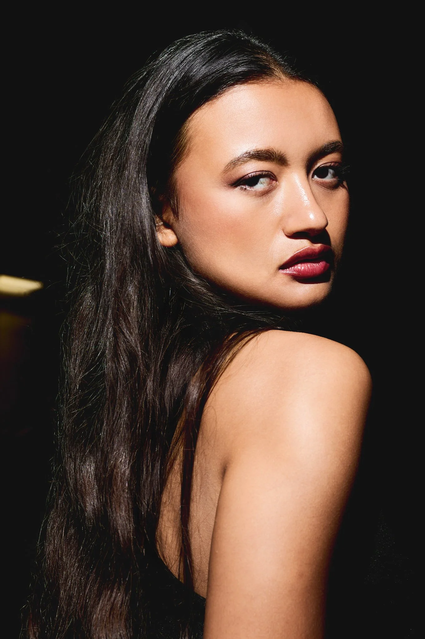 Close-up of a woman with long dark hair, wearing makeup, looking over her shoulder against a dark background.
