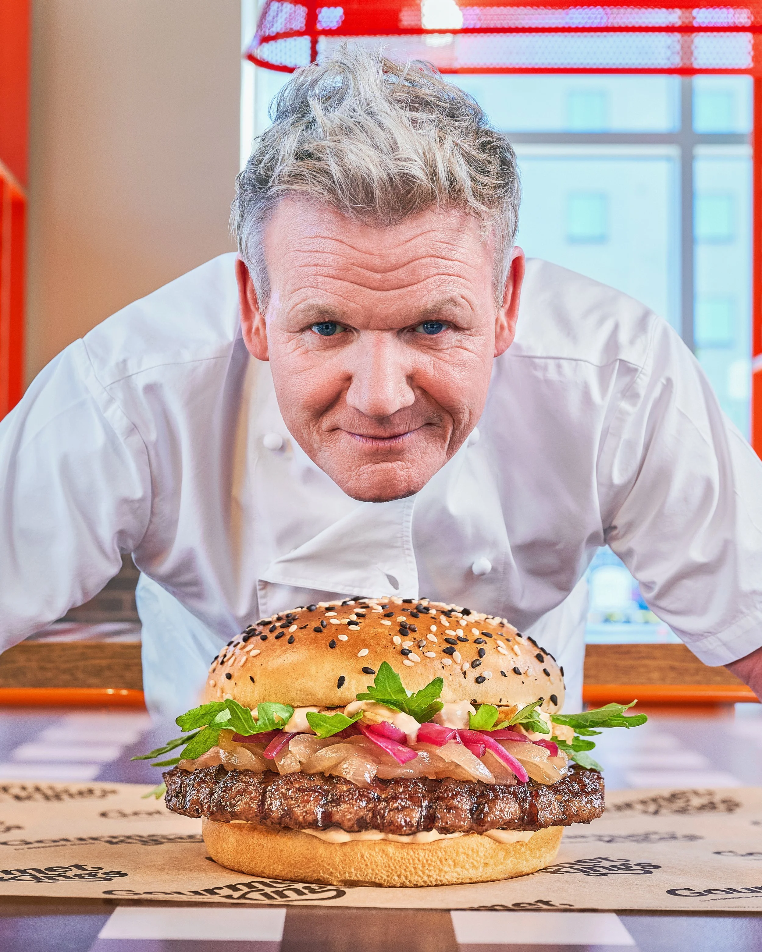 A chef crouching in front of a large burger on a wooden surface.