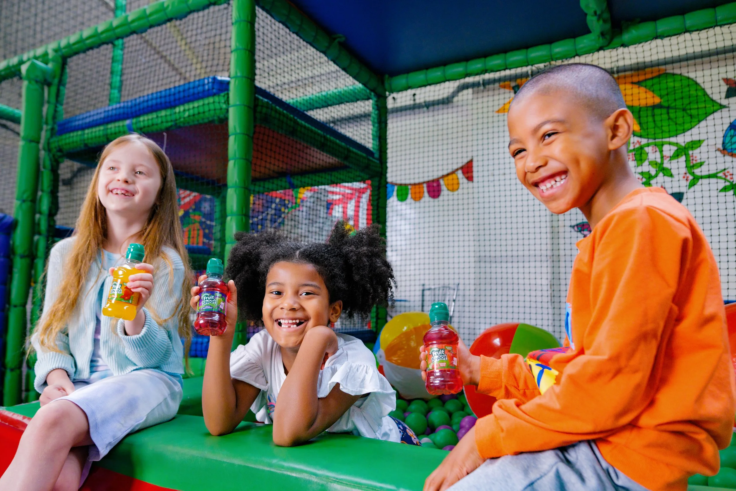 Three children, two girls and a boy, sitting on a padded surface in an indoor play area with colorful ball pit balls and netted climbing structures around them, holding bottles of juice and smiling.