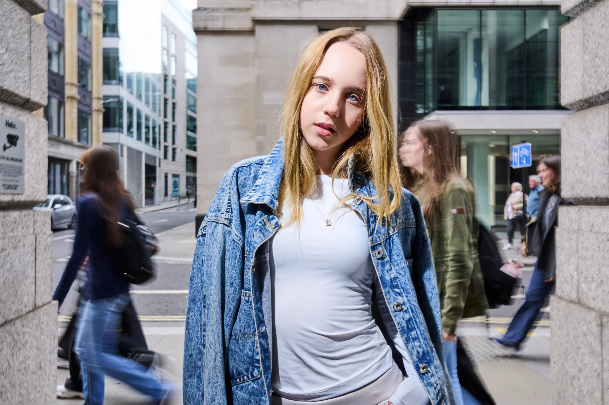 A young woman with blonde hair in a denim jacket and white shirt standing in an urban setting with people walking in the background.