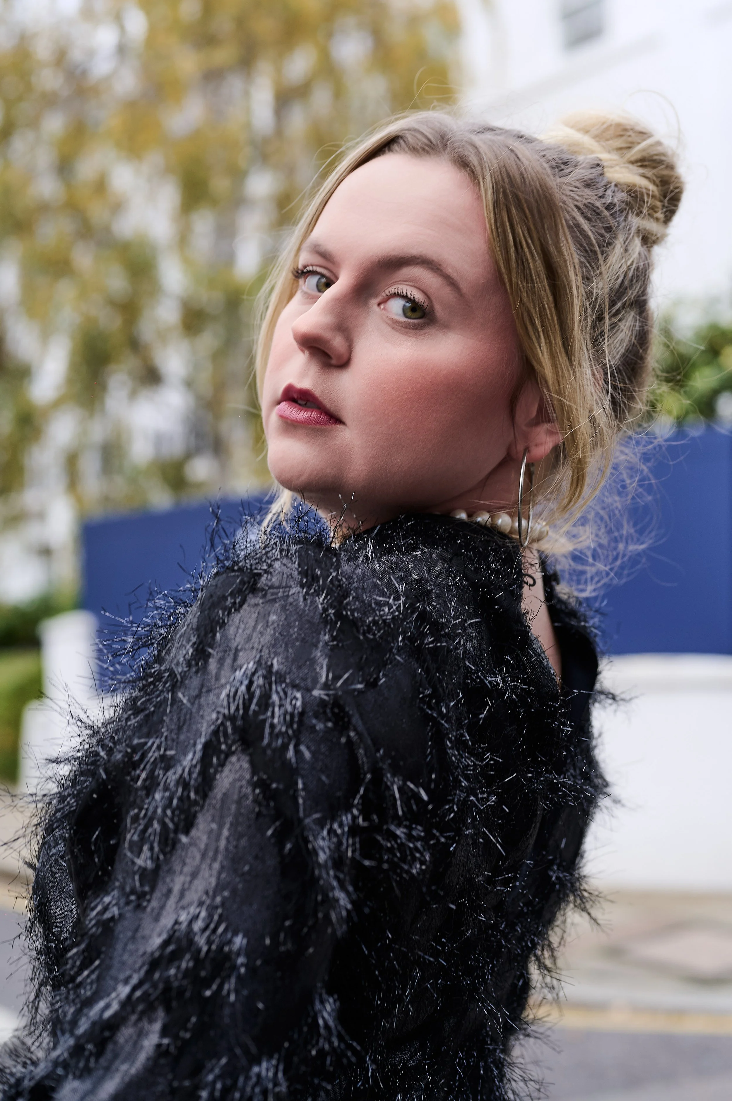 A woman with blonde hair styled in an updo, wearing makeup and a black, textured dress with feather-like details, standing outdoors with trees in the background.