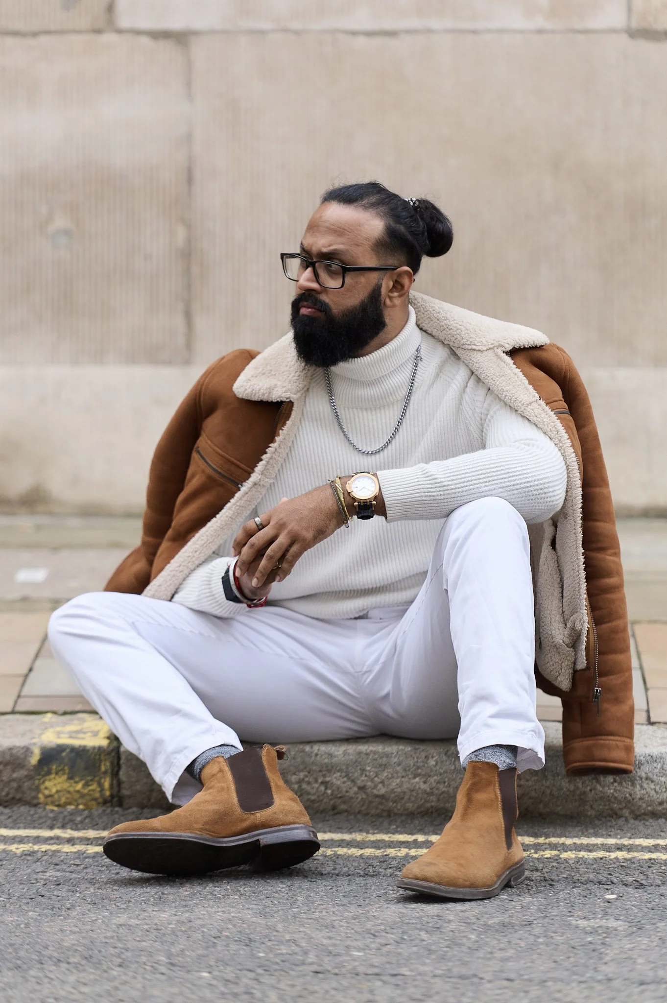 A man with glasses, a beard, and long hair in a bun sitting on a curb in front of a beige wall. He is wearing a white turtleneck sweater, white pants, a brown jacket, and brown boots, with a watch and bracelets on his left wrist and a chain necklace.