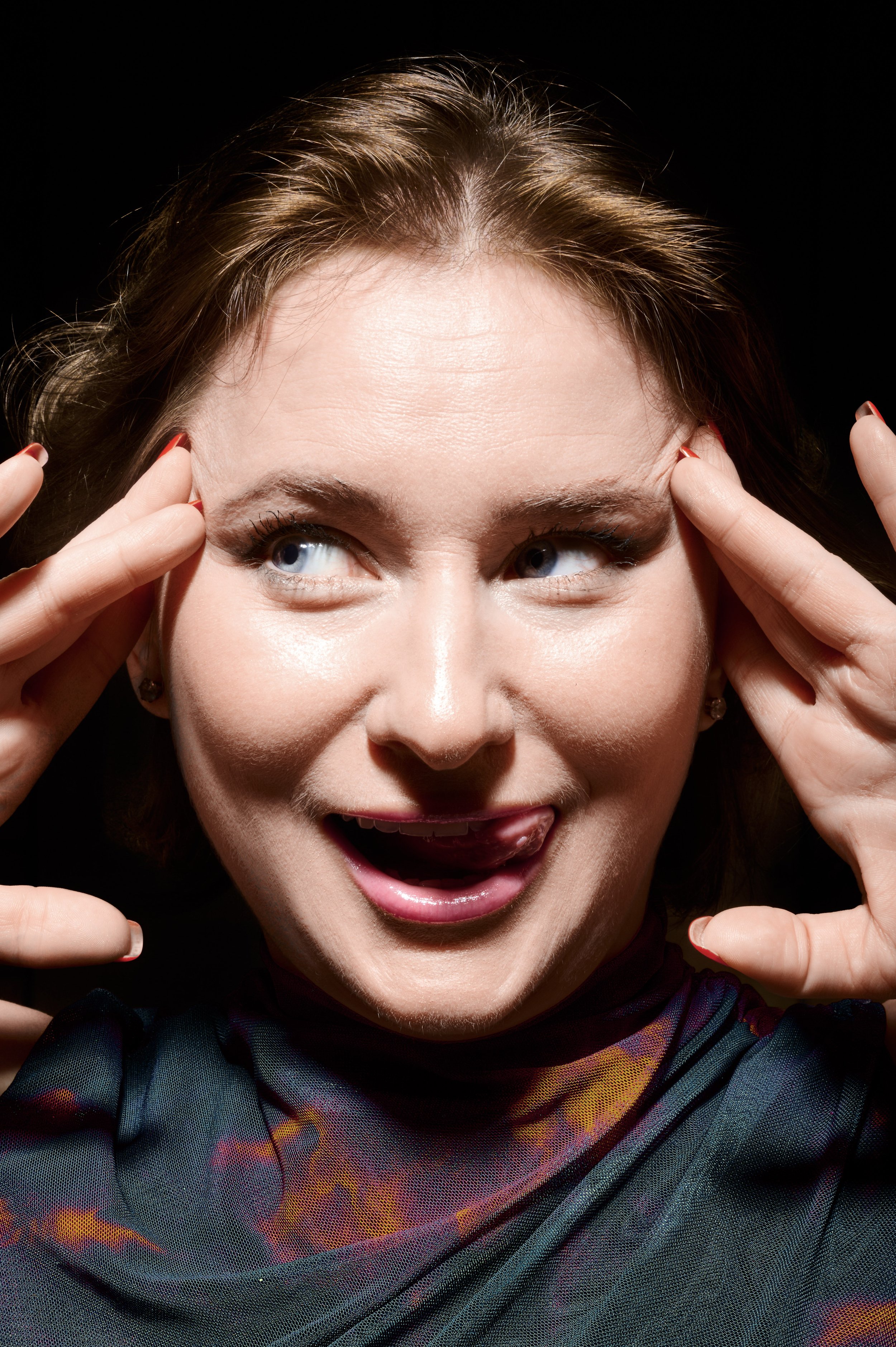 A woman with brown hair touching her forehead with both hands, smiling playfully with her tongue slightly out, against a black background.