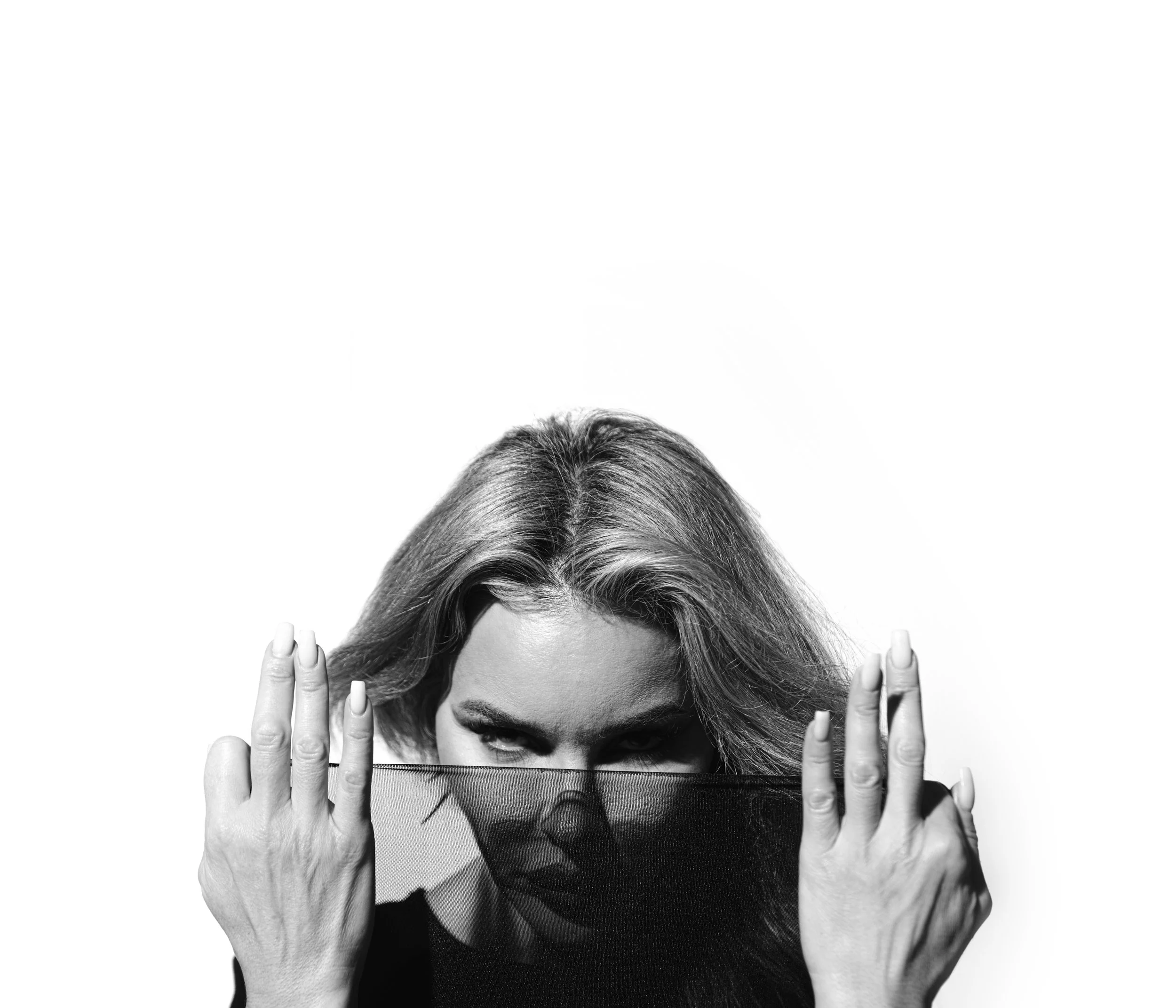 Black and white photo of a woman with light hair holding a patterned cloth with her eyes peeking over it, against a white background.