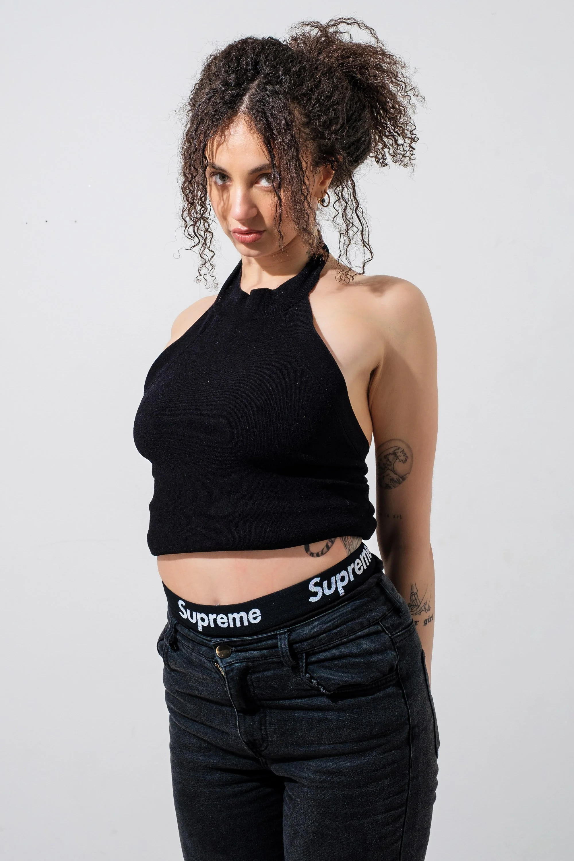 Young woman with curly hair wearing a black crop top and black Supreme waistband standing against a white background.