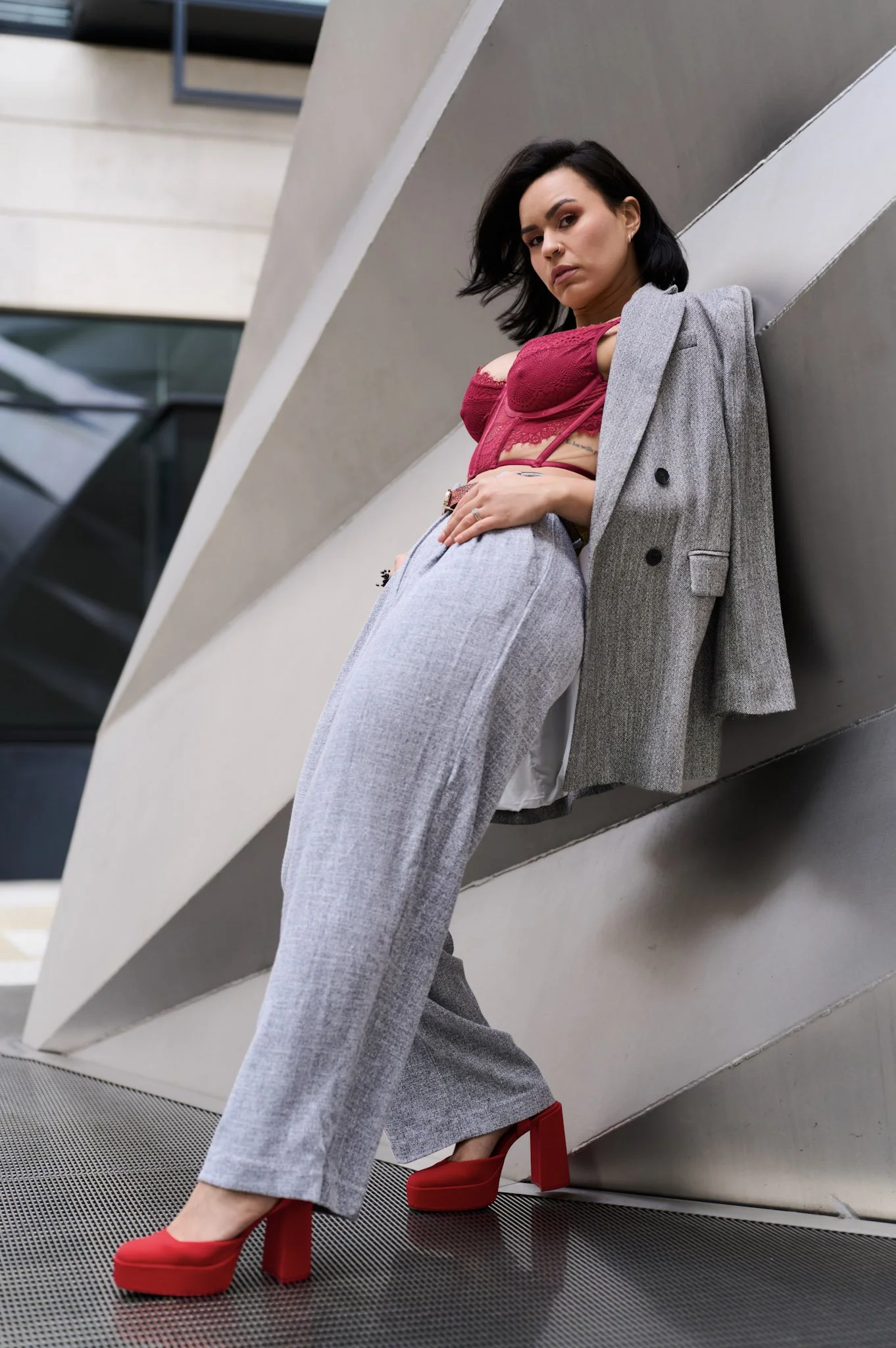 A woman with dark hair, wearing a red lace top, gray high-waisted pants, a gray blazer draped over her shoulders, and red high heels, leaning against a modern architectural wall.