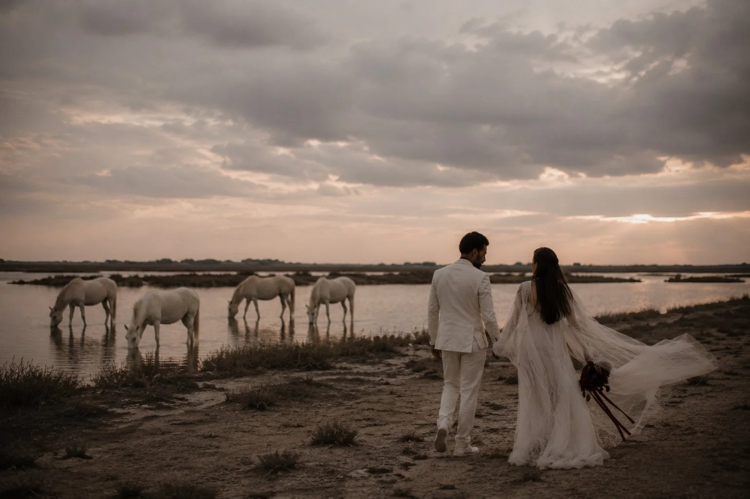 Bride and groom walking through the Camargue at sunset beside white horses during a luxury destination wedding in France.