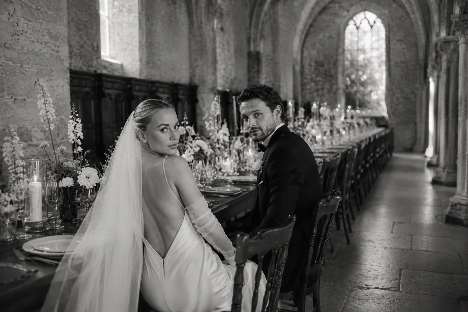 Bride and groom seated at an elegant candlelit reception table inside Abbaye des Vaux de Cernay, both looking toward the camera in a refined editorial wedding setting.