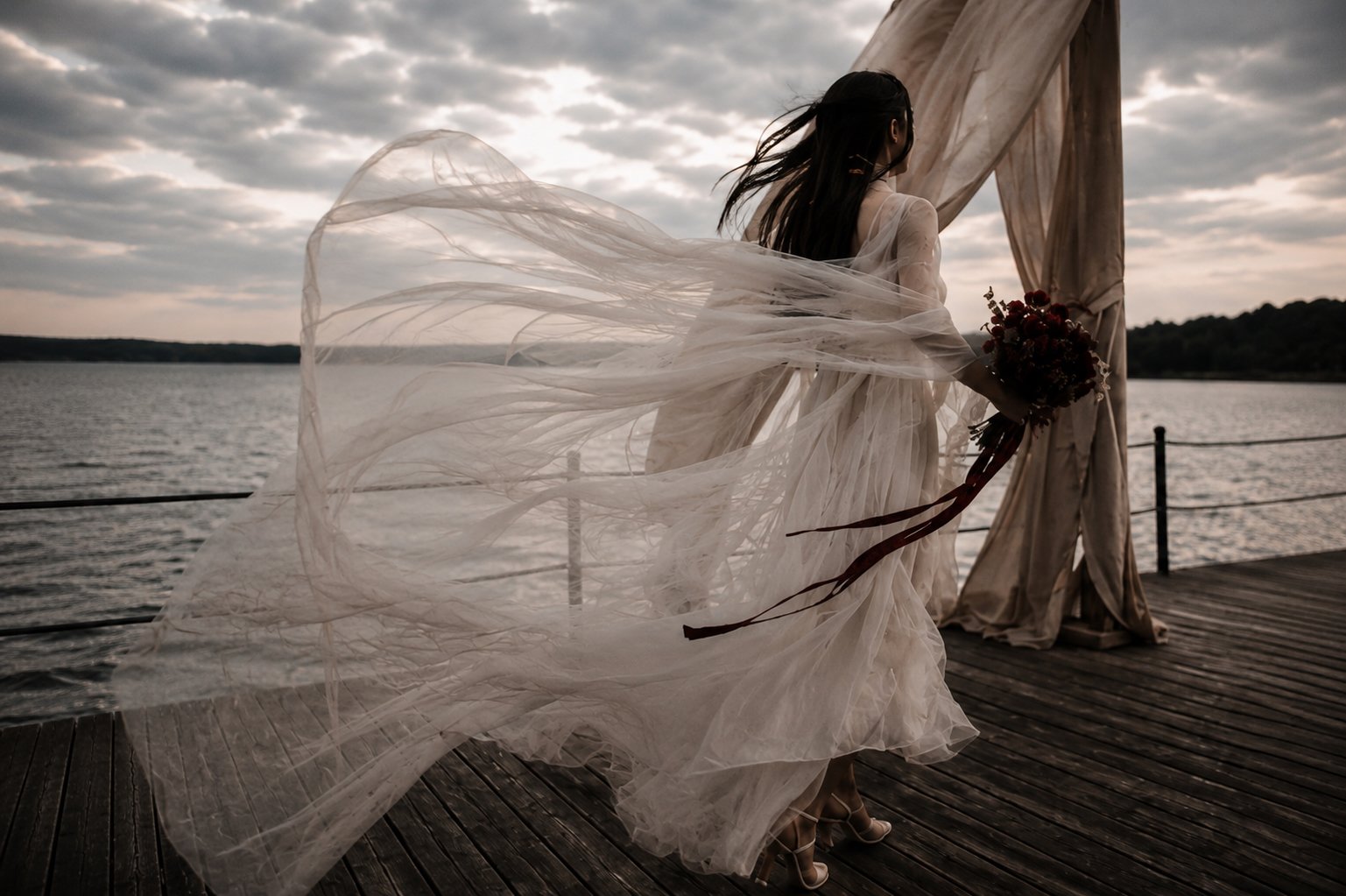 Editorial portrait of a bride  with a couture tulle gown and a  long flowing veil and burgundy bouquet beside a lakeside ceremony in the Camargue. Fine art destination wedding photography in France.