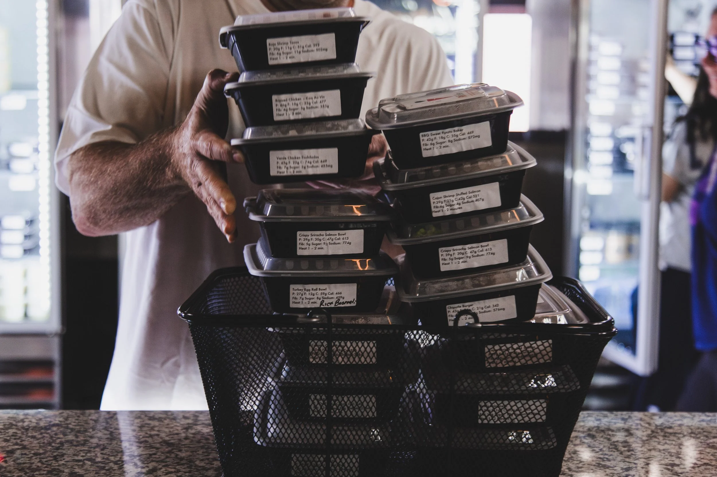 Person holding a stack of prepared food containers above a black wire basket on a countertop.