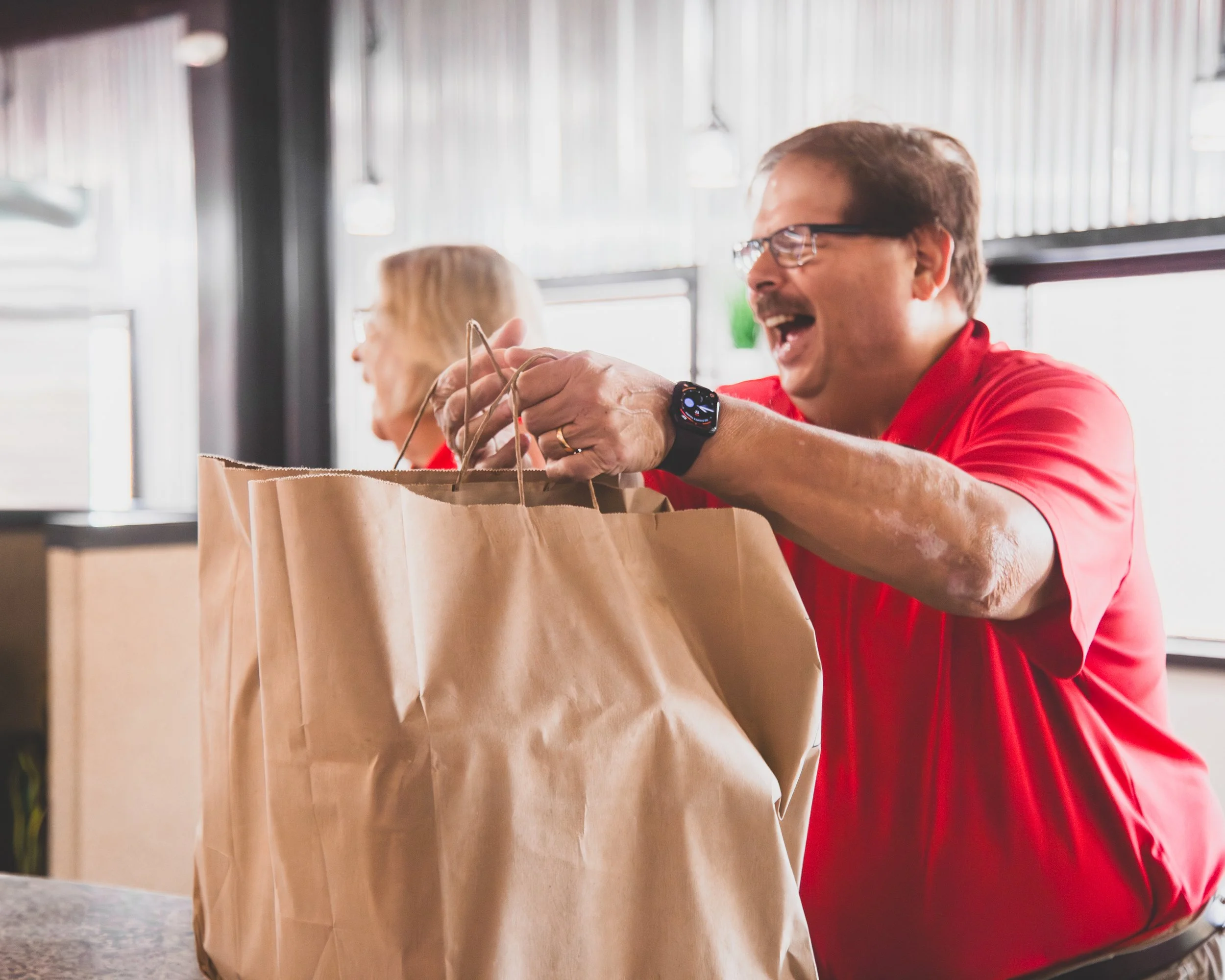 A man wearing glasses and a red shirt joyfully reaches into a large brown paper bag, with another woman in the background at a cafe or restaurant.