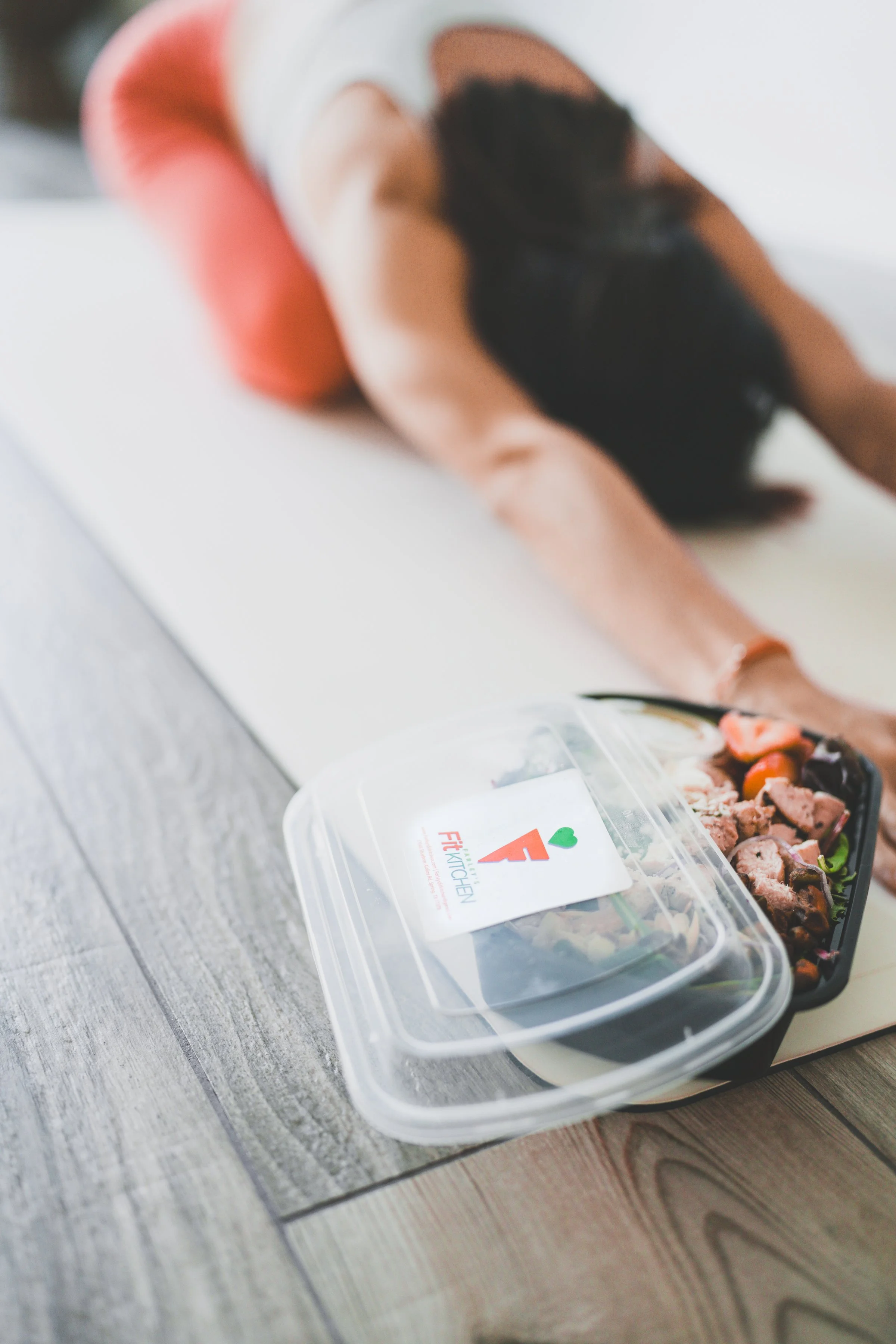 A woman lying on a yoga mat doing a stretch. In the foreground, there is a container of salad with a label that reads 'Fit Kitchen' placed on a wooden floor.