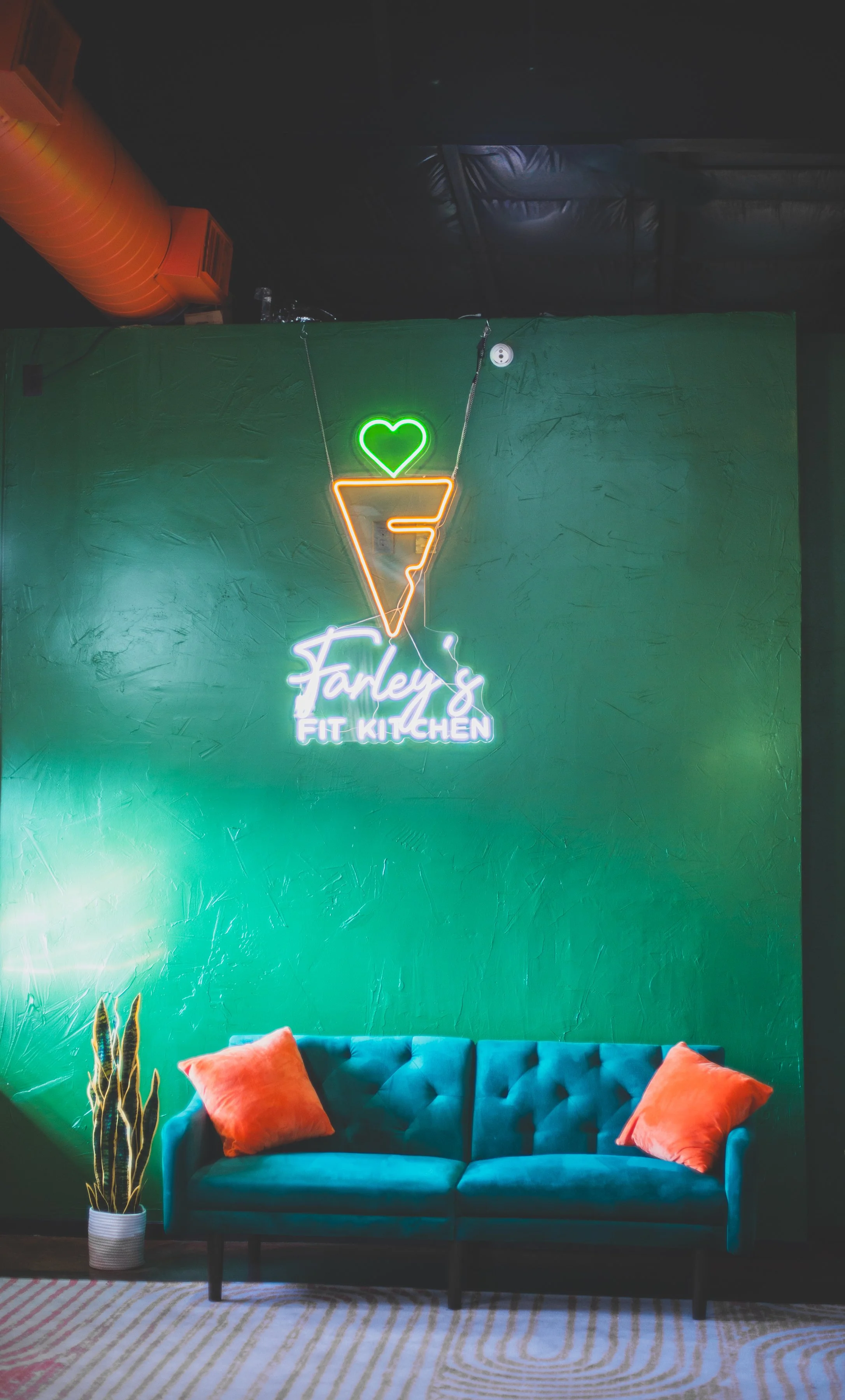 A neon sign reading 'Farley's Fit Kitchen' with an ice cream cone and a green heart above it, hanging on a green wall above a blue sofa with two orange pillows, and a tall plant to the left.