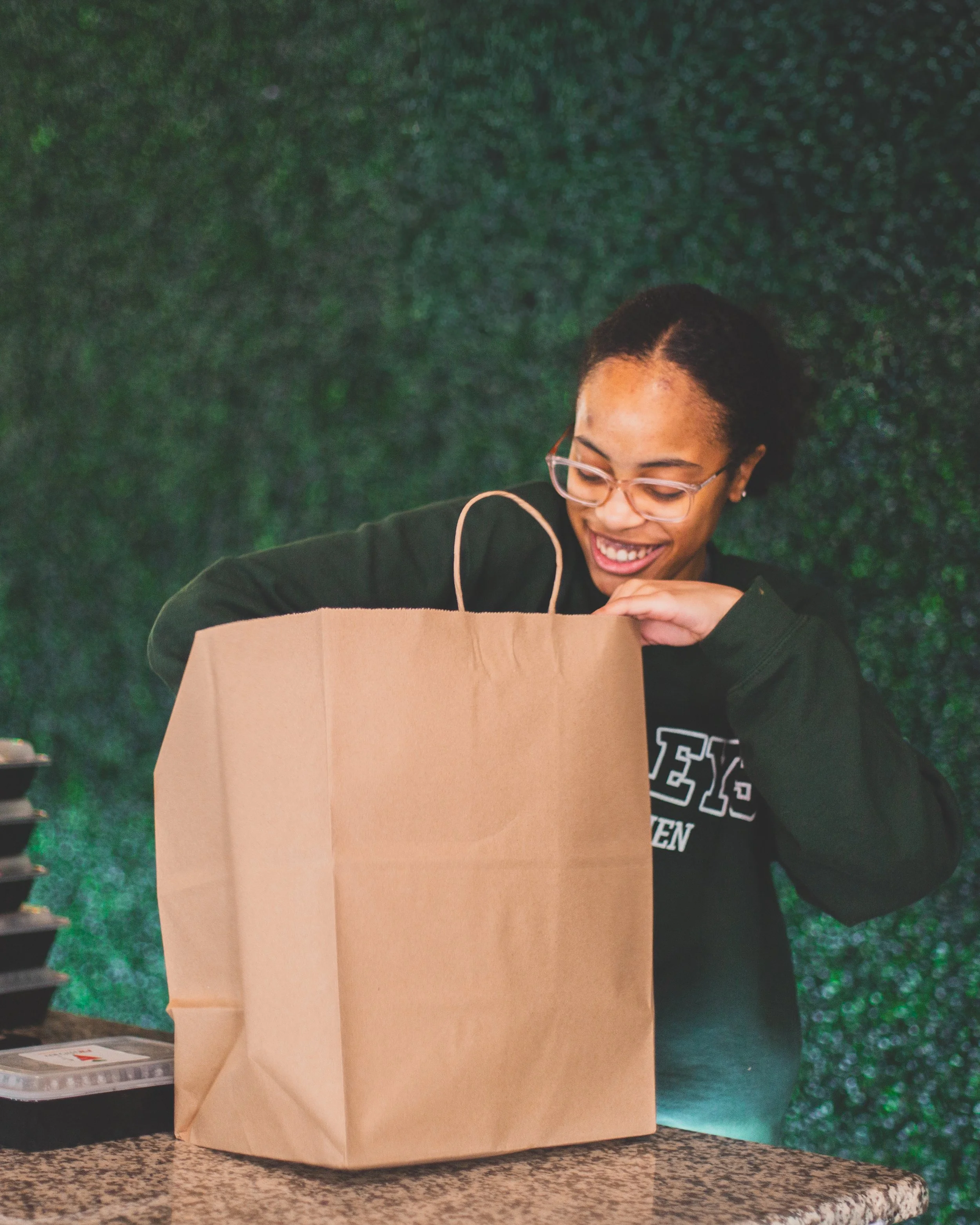 A woman wearing glasses and a black sweatshirt is smiling as she looks into a large brown paper bag on a counter, with a green textured background behind her.