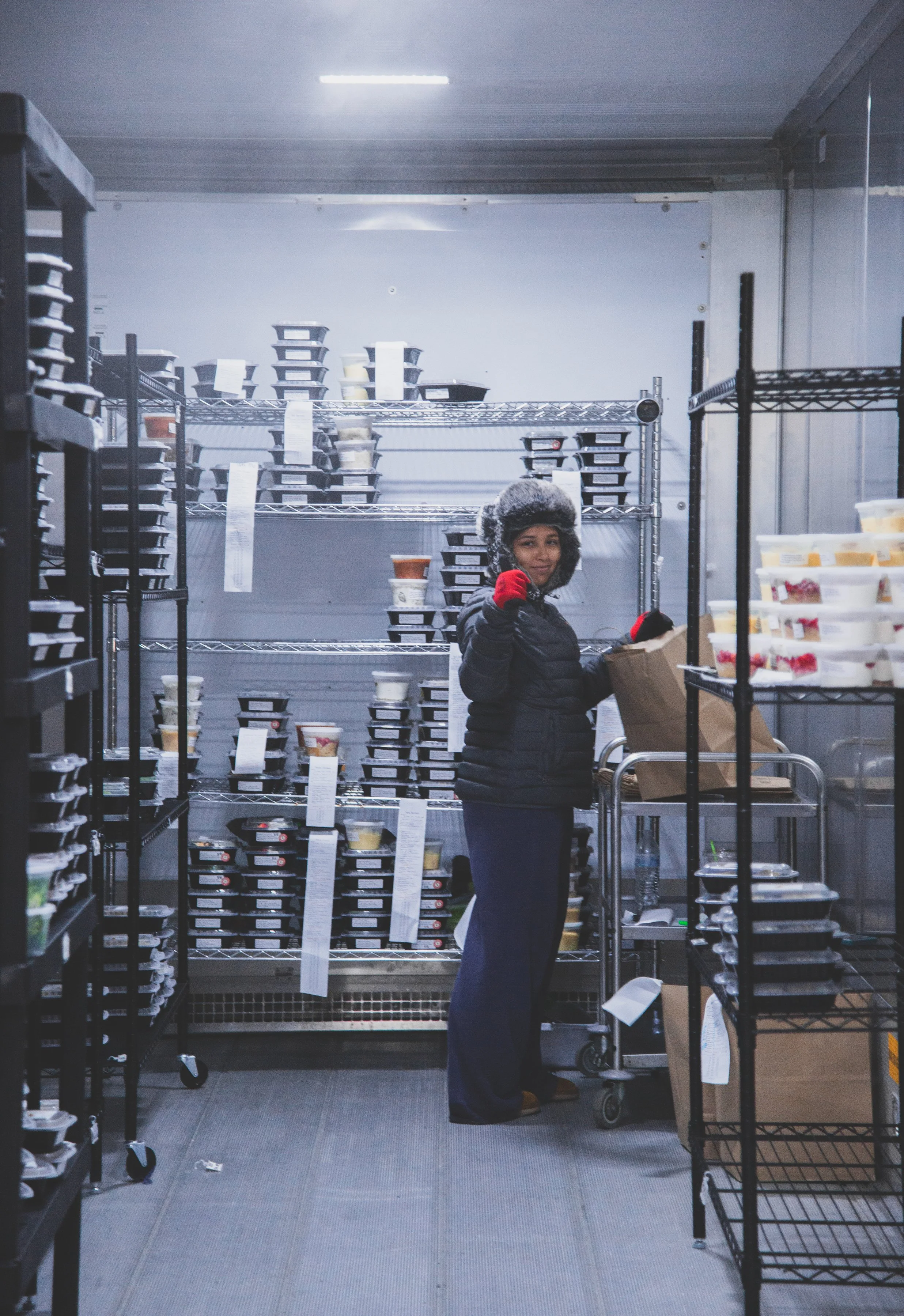 Woman in a winter jacket and furry hat in a walk-in refrigerated storage room, holding an open brown paper bag and giving a thumbs-up.