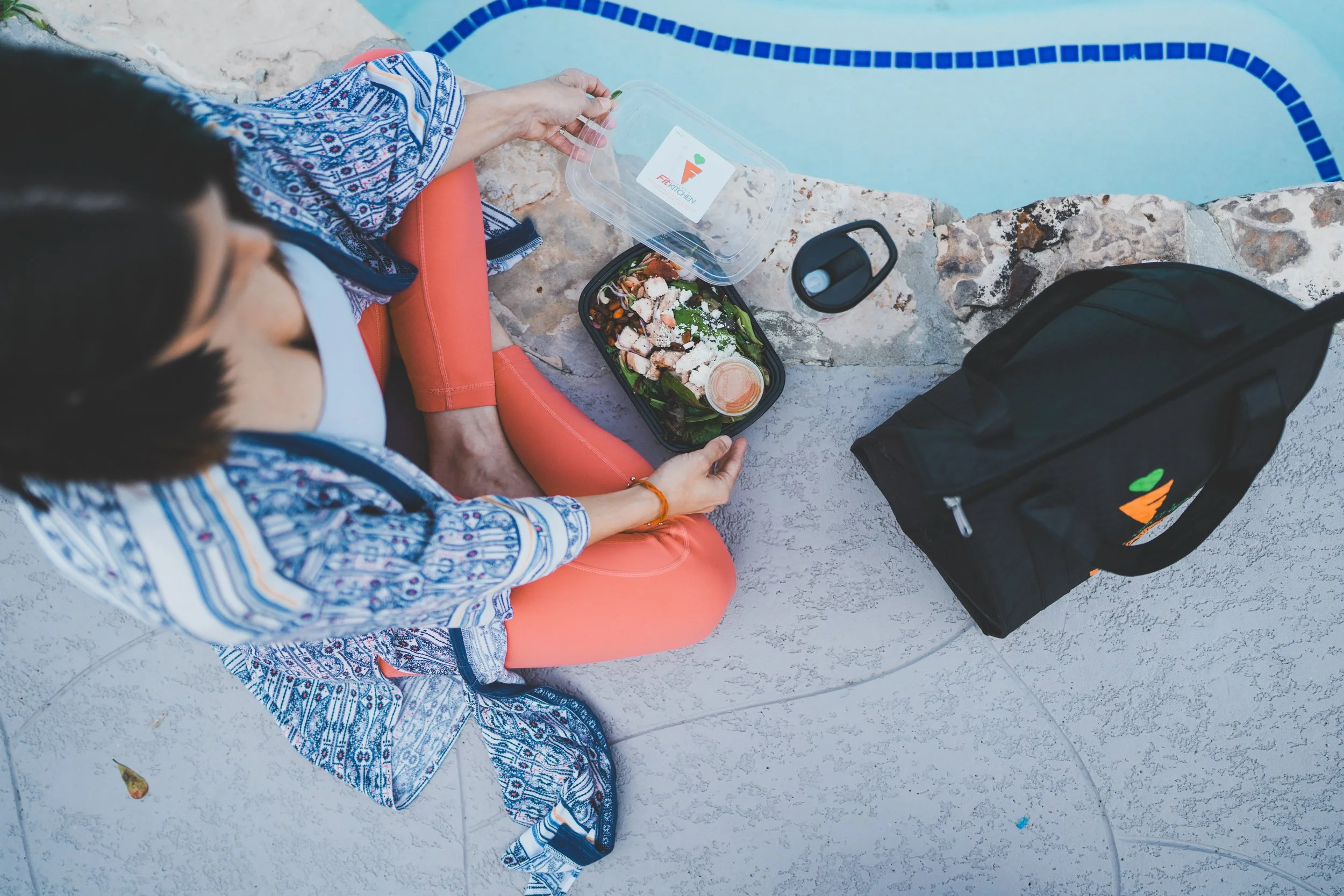 A person sitting by a pool with a salad container, a drinking cup, and a black bag with a logo on a stone ledge.
